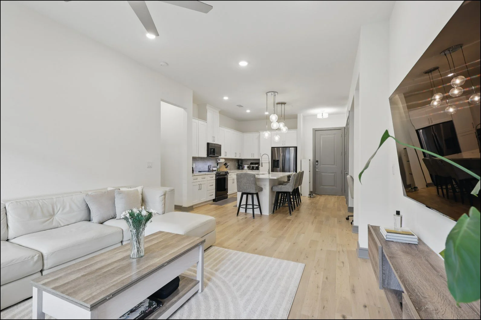 Modern open-plan living room and kitchen with light wood flooring, white cabinetry, gray seating, and contemporary lighting fixtures.