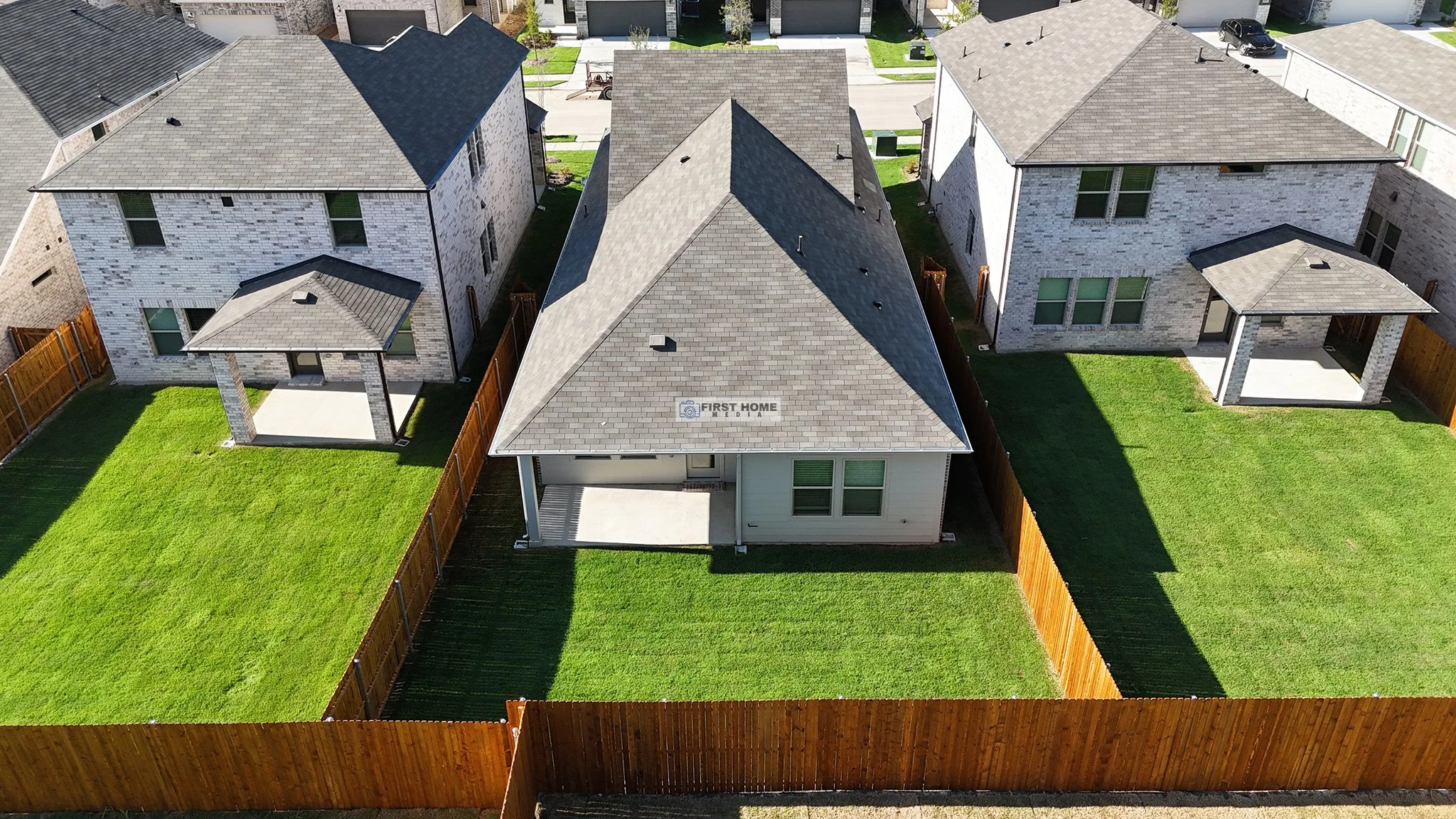 An aerial view of three neighboring houses with well-maintained green lawns, grey shingle roofs, and wooden fences, in a suburban neighborhood.