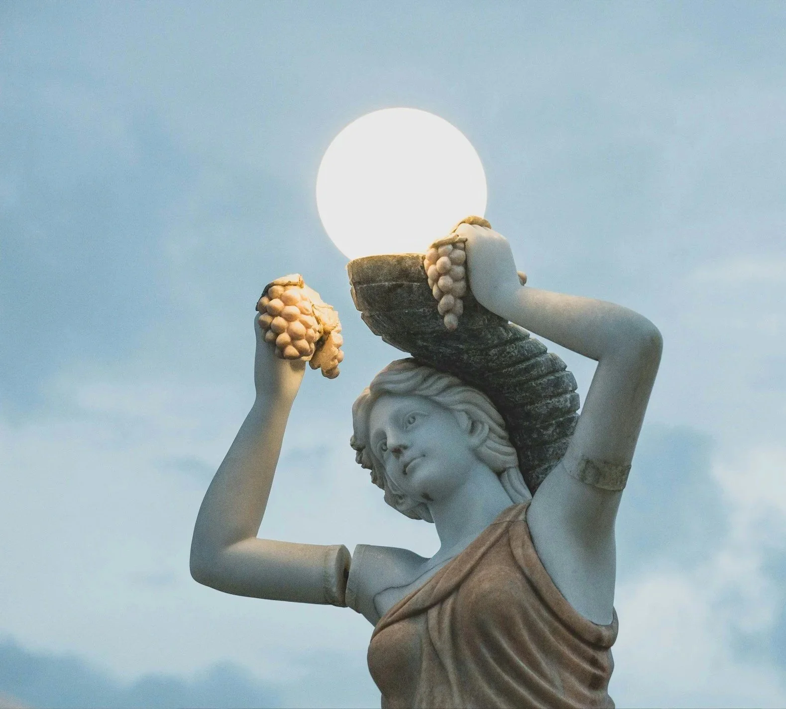 Close-up of an archetypal sculpture of a woman holding a large object that resembles a sun or moon, against a backdrop of the sky with the sun.