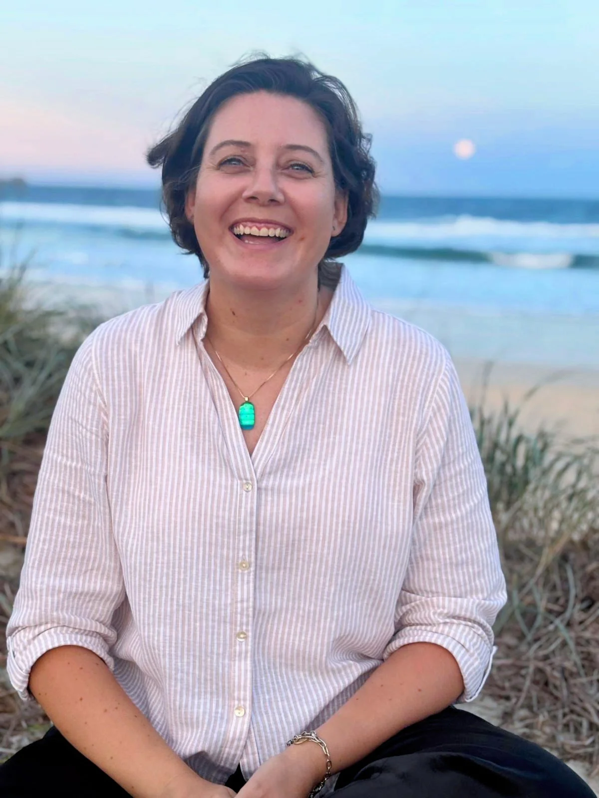 Jule Carter from Securely You counselling, with short dark hair smiling on a beach during sunset, wearing a light pink striped button-up shirt and a turquoise necklace.