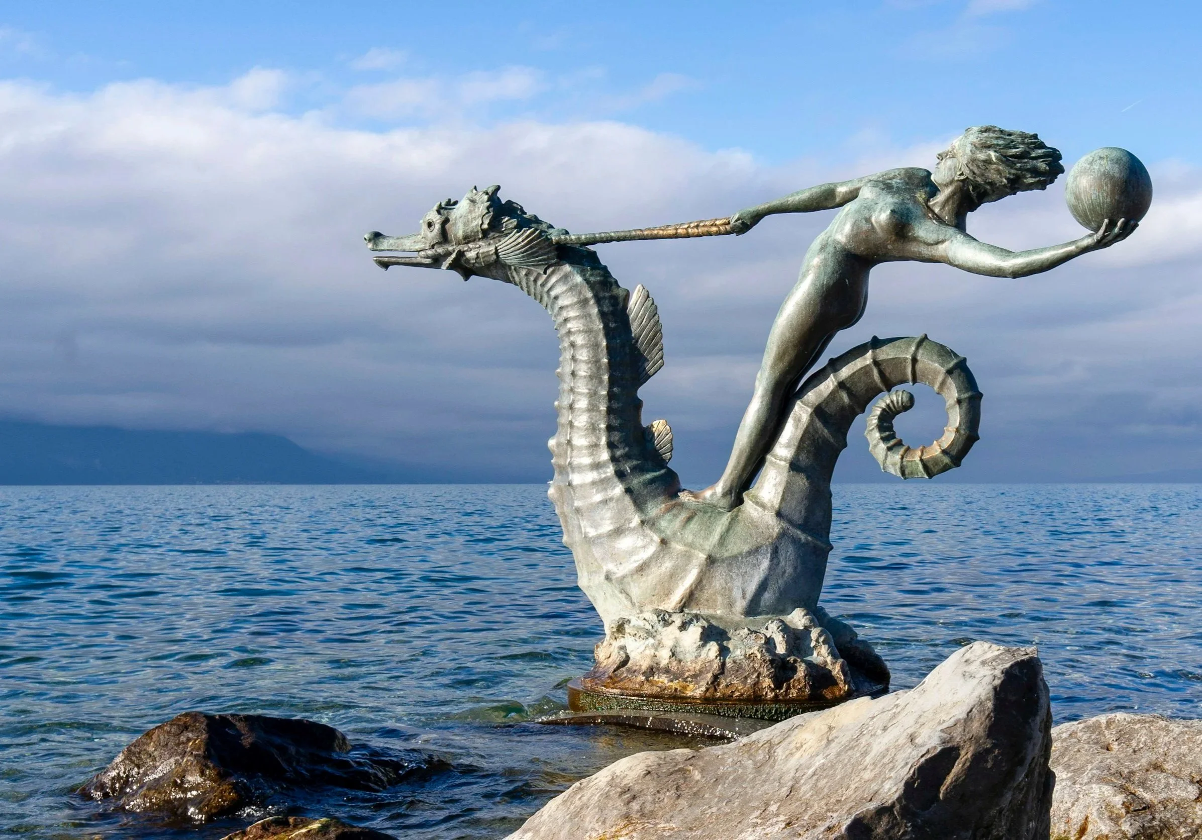 A sculpture of a mermaid with flowing hair holding a ball in her right hand and grasping a seahorse with her left hand, positioned on a rock by the water under a partly cloudy sky.