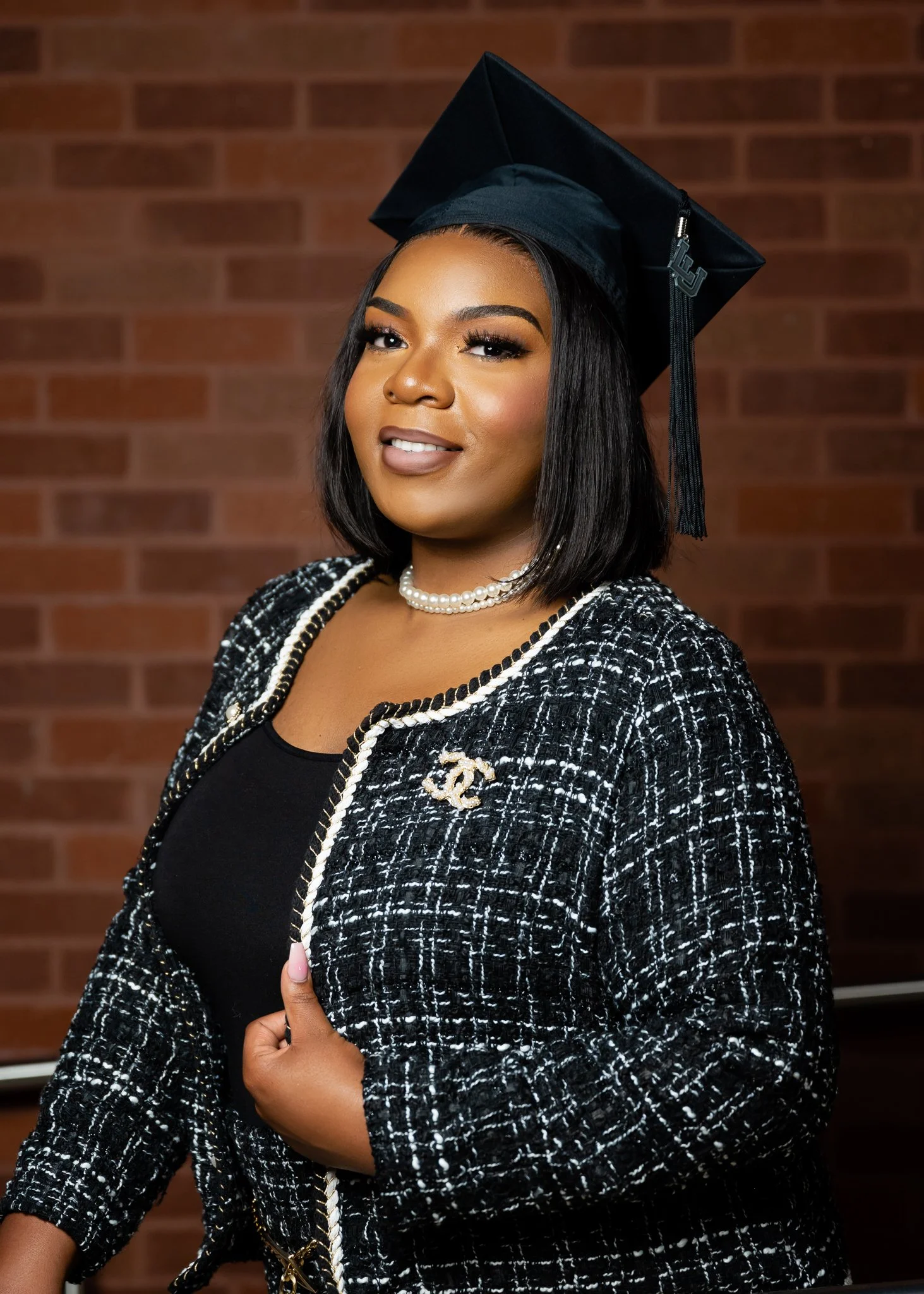 A woman in a graduation cap and black-and-white tweed jacket, standing against a brick wall.