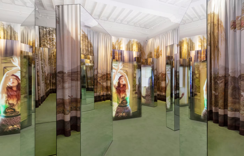 An art installation features multiple mirrored pillars and curtains with a nature scene and a woman with red hair on them.