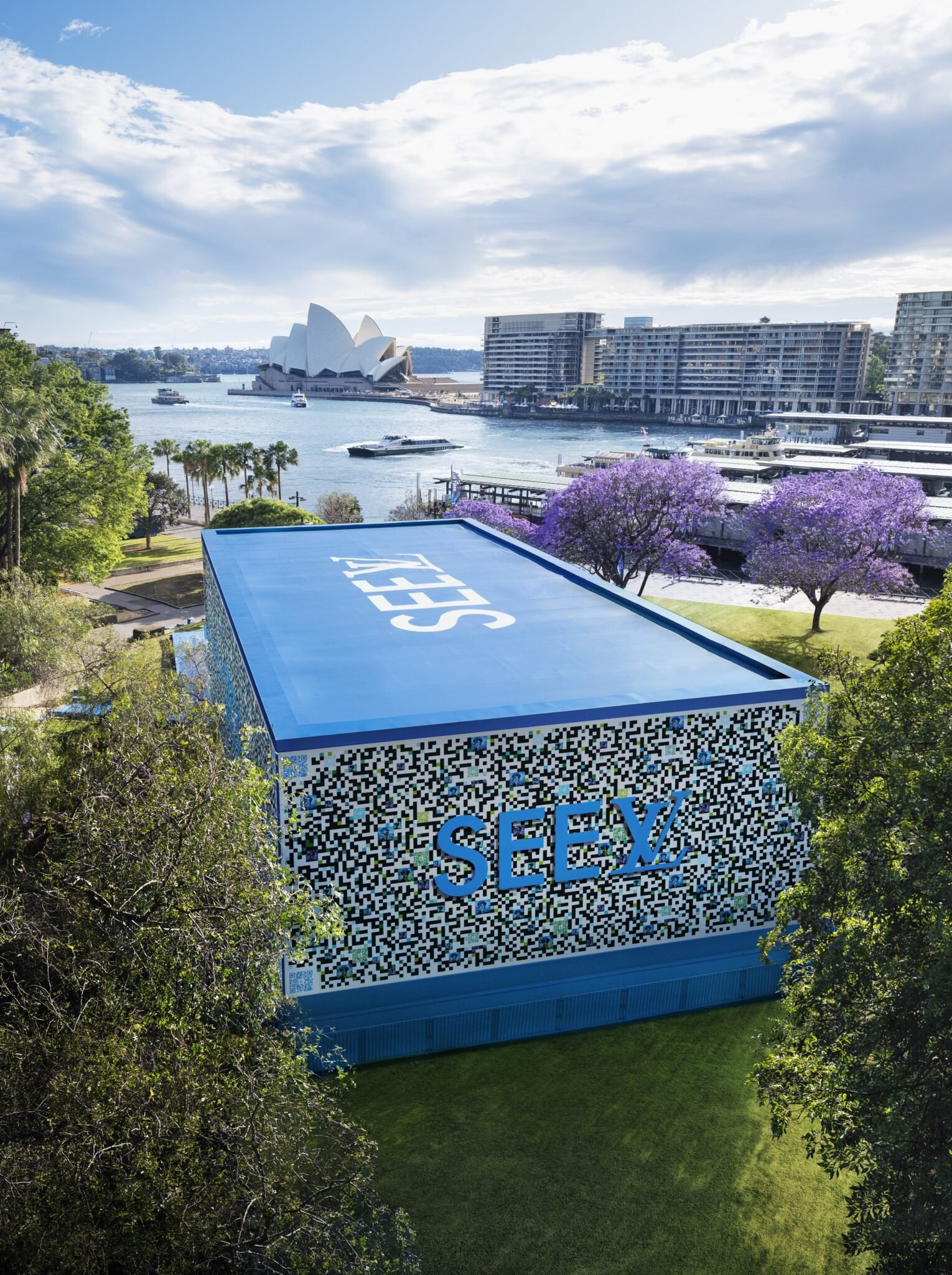 A large QR code-shaped structure with the word 'SEEX' on it, situated in a park near the waterfront with trees and purple flowering trees. In the background, there is a view of the Sydney Opera House, boats on the water, and modern buildings, indicating a cityscape environment.
