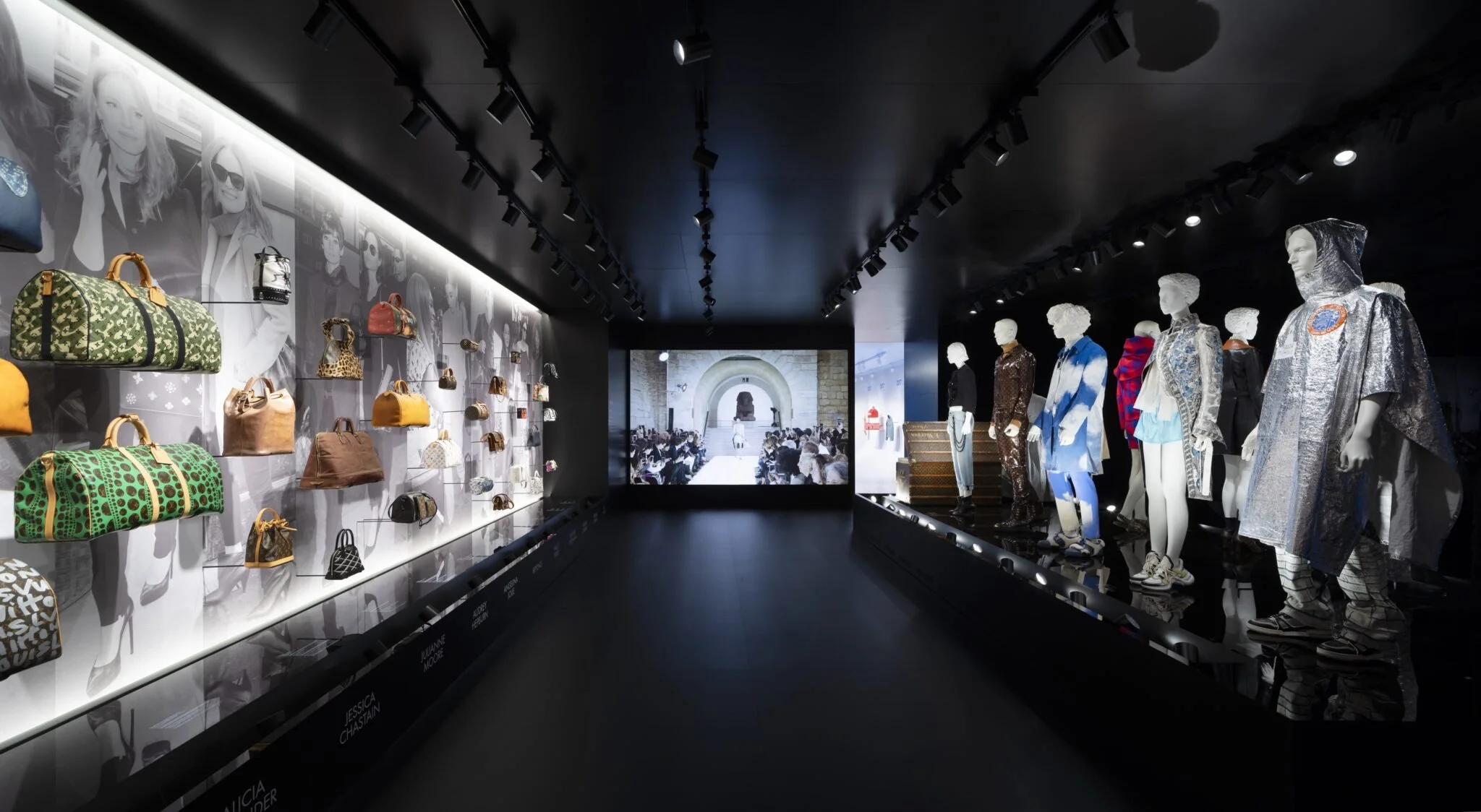 Museum exhibit displaying designer handbags on the left wall and mannequins dressed in fashion on the right, with a large digital screen in the background showing an audience and a historic building.