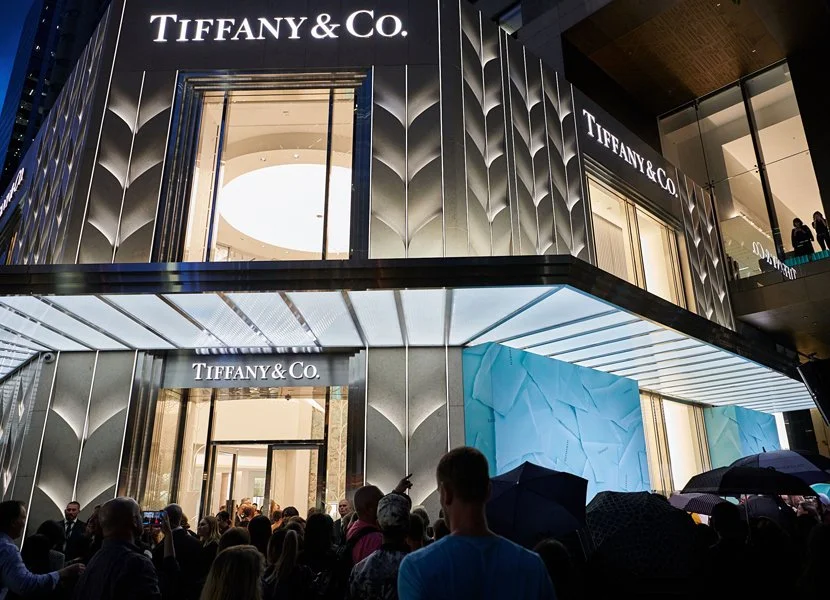 Exterior view of a Tiffany & Co. store with a crowd of people outside, some with umbrellas, on a rainy day. The store has a modern design with metallic panels.