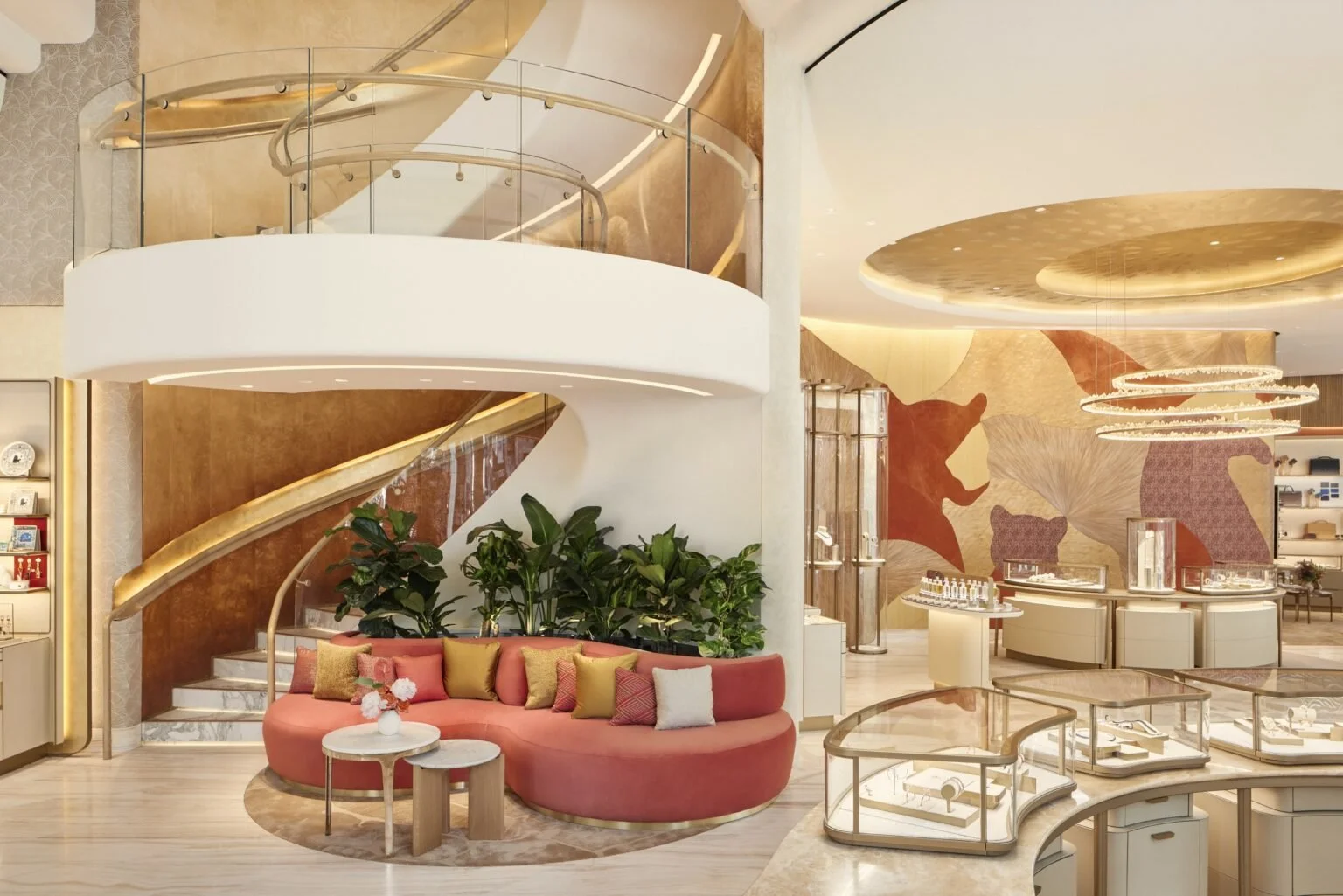 Interior of a modern, luxurious store or hotel lobby with a curved pink sofa, colorful pillows, green plants, marble staircase, glass railings, and decorative ceiling lights.