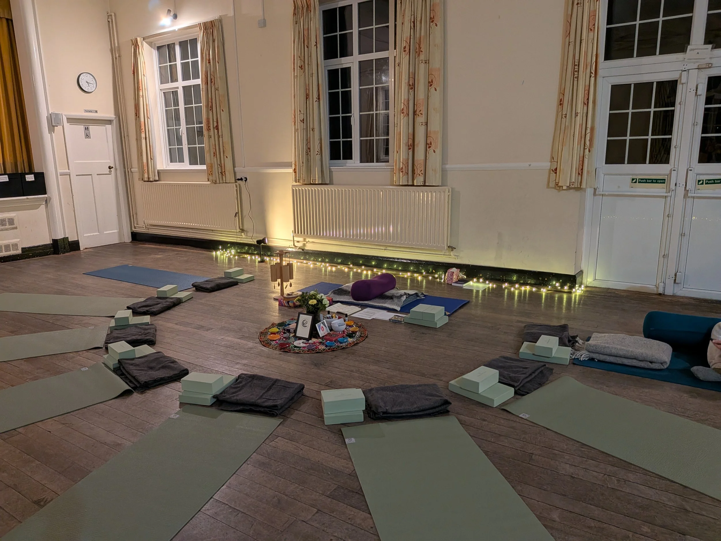 Green mats and grey blankets layed out on the floor with a colourful circular piece of fabric in the middle with a vase of flowers on, ready for the yoga nidra event in Clifton, Bedforshire.