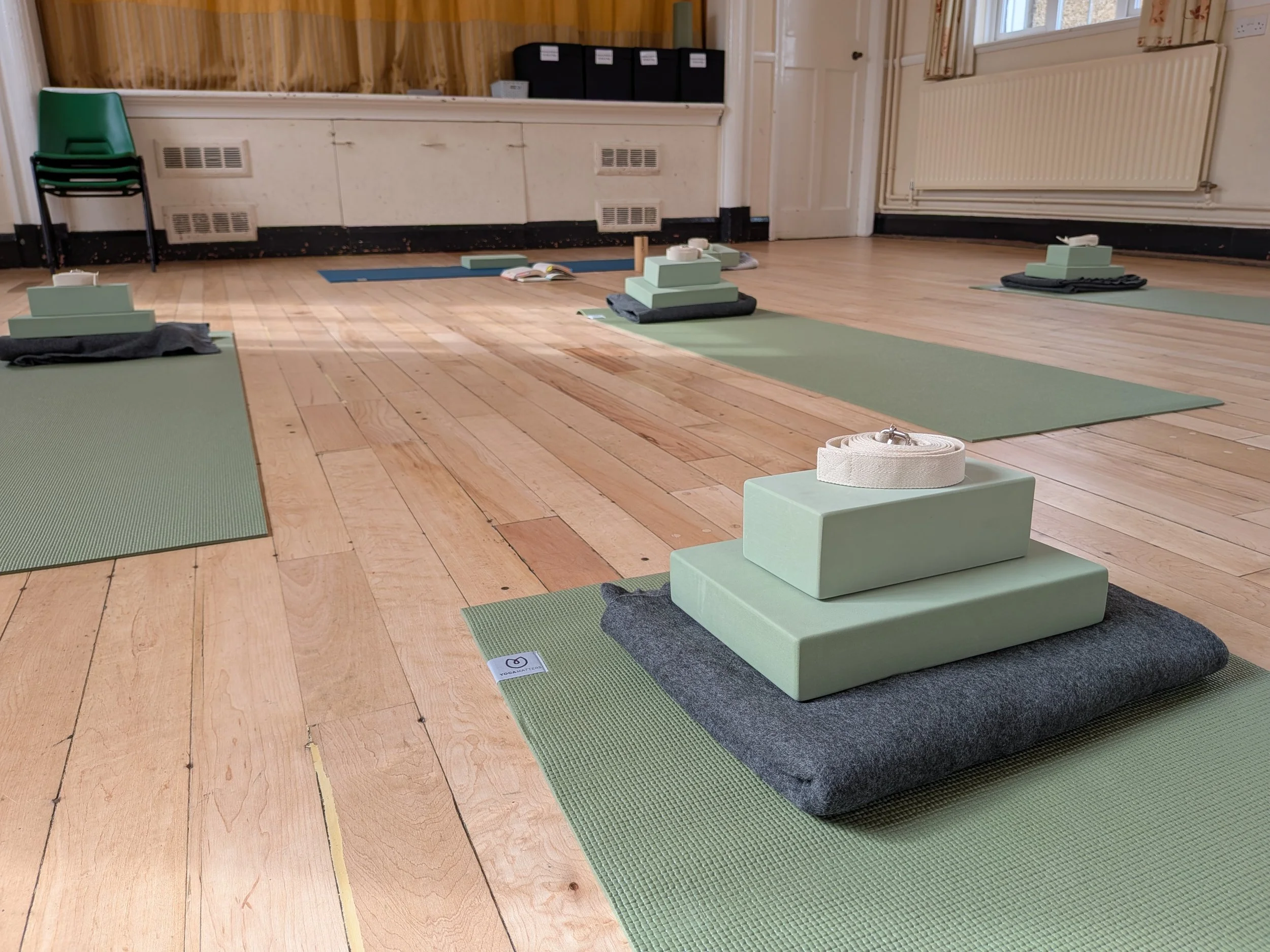Green yoga mats with blankets, blocks, bricks and yoga straps layed out on the floor in a bright and spacious hall ready for a yoga class in Clifton, Bedfordshire