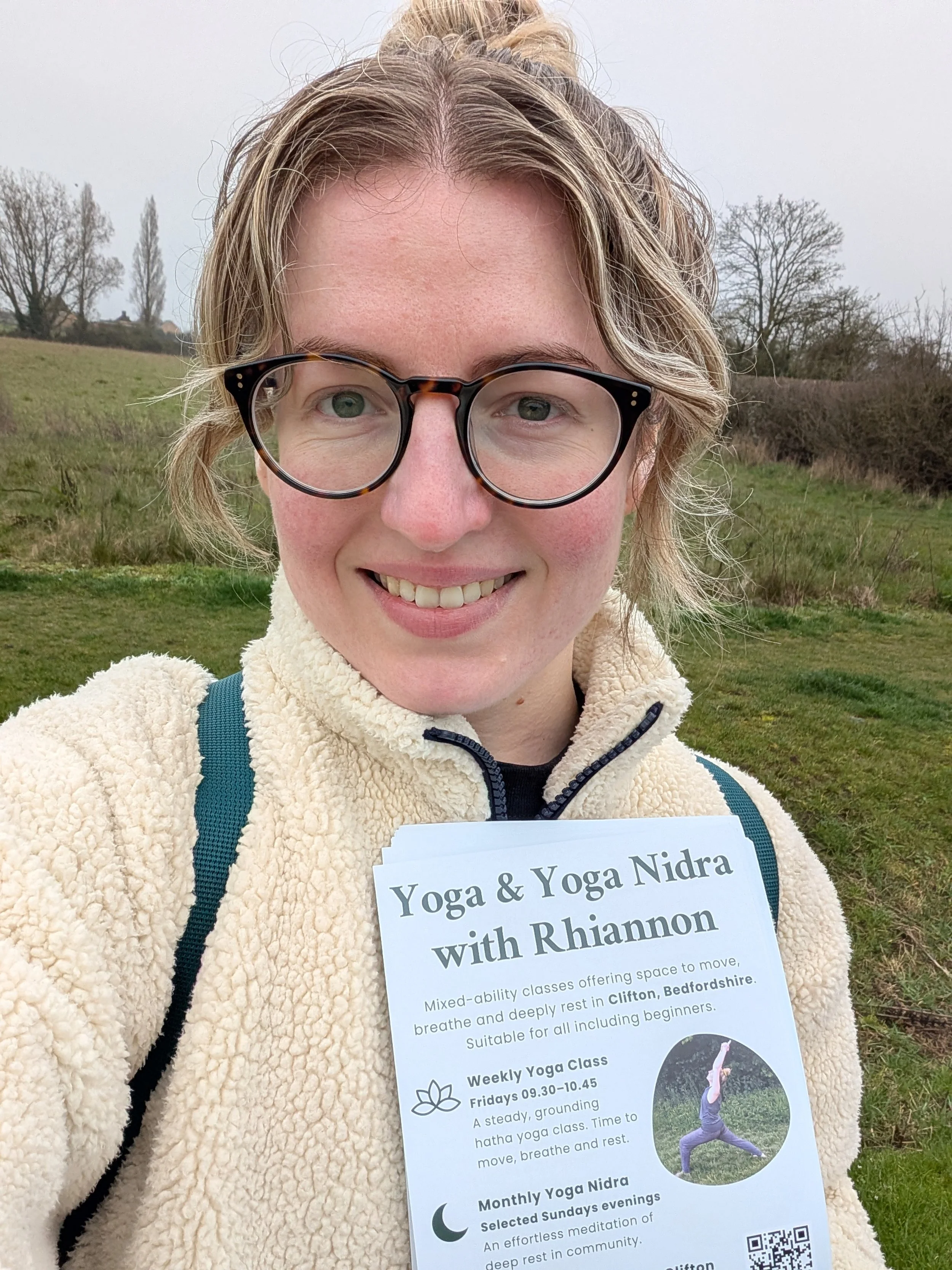 A woman wearing glasses smiling at the camera, standing outside and holding leaflets about yoga class and yoga nidra in Clifton, Bedfordshire.