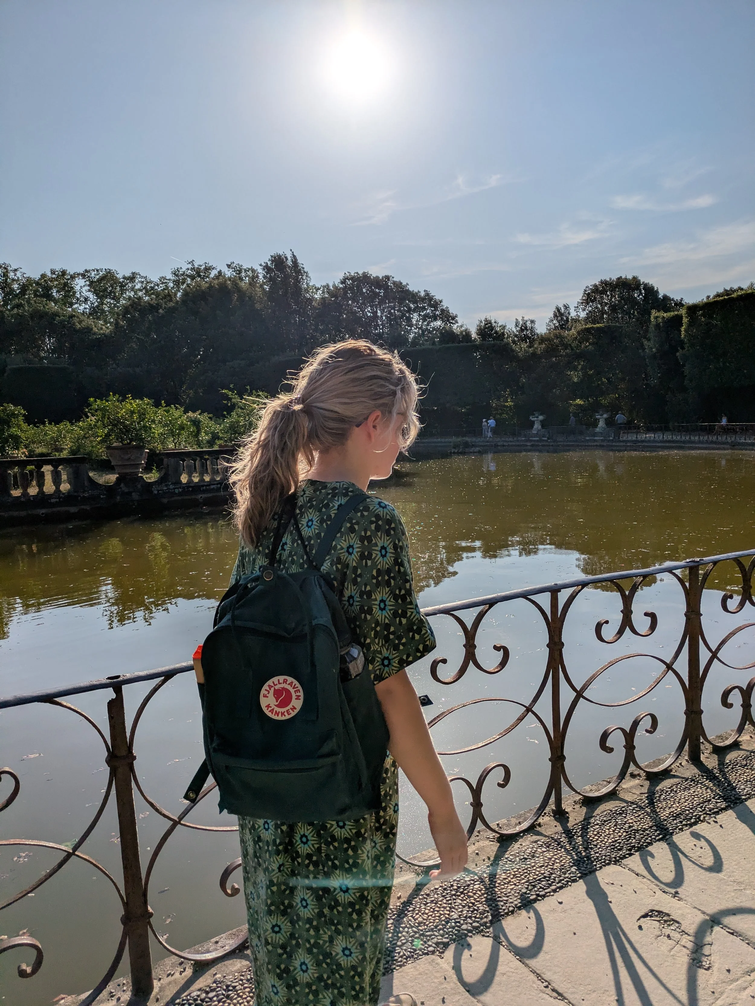 A woman walking next to a lake carrying a green rucksack and blonde hair in a ponytail with the sun shining.