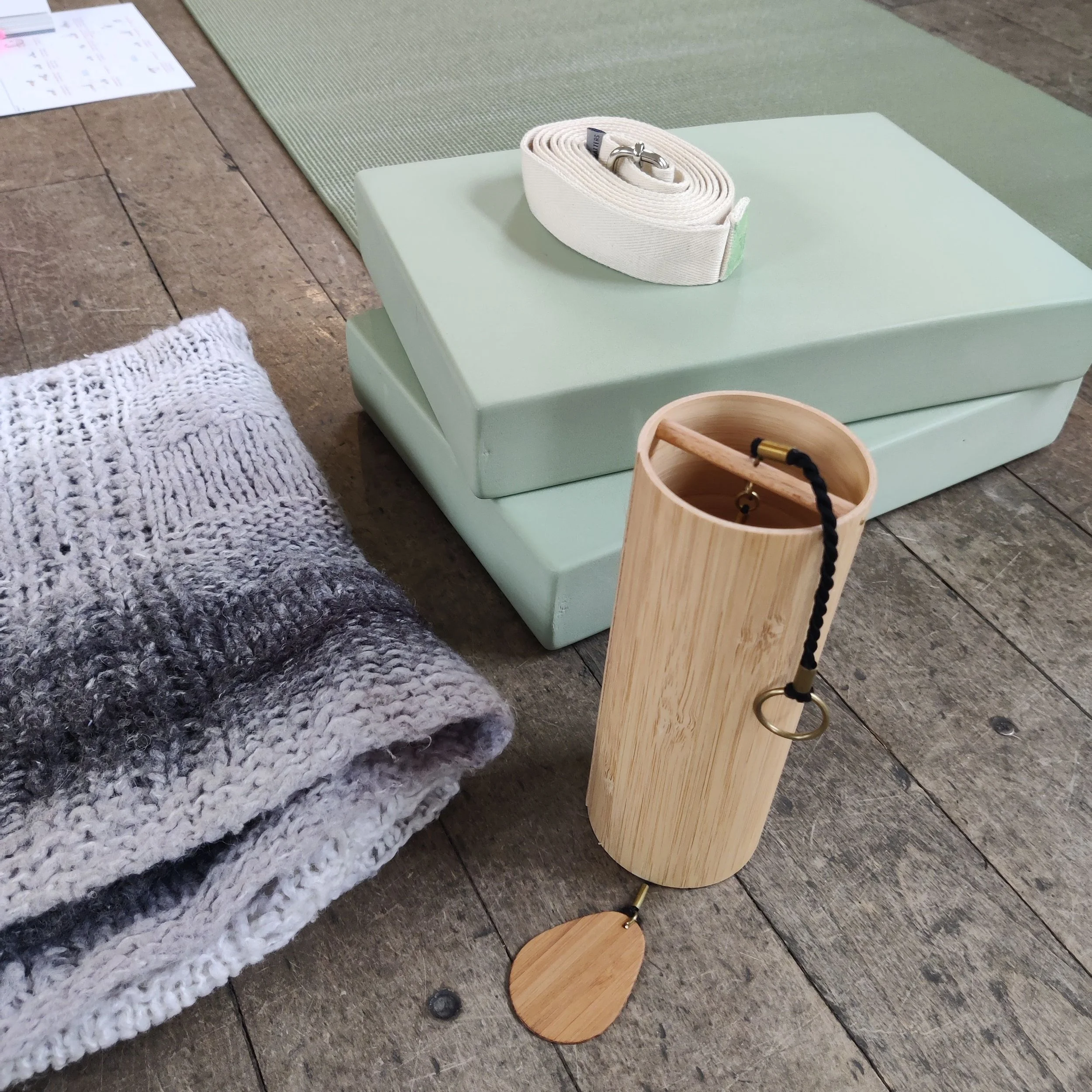 Green yoga blocks, grey blanket, yoga strap and koshi chime on wooden floor boards..