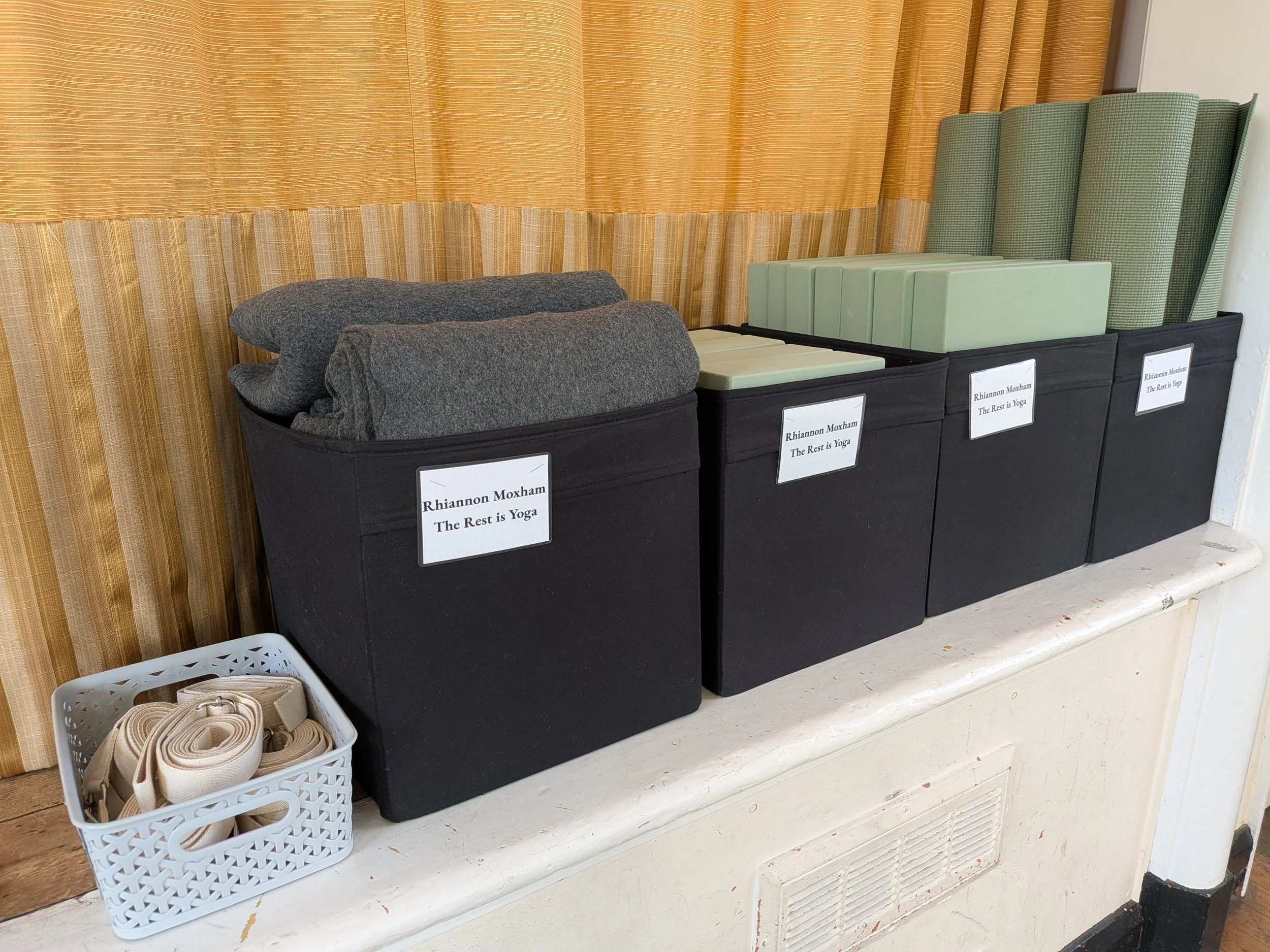 Green yoga mats, blocks, bricks and blankets in black storage boxes and a small grey box of yoga straps ready for use at yoga class in Clifton, Bedfordshire