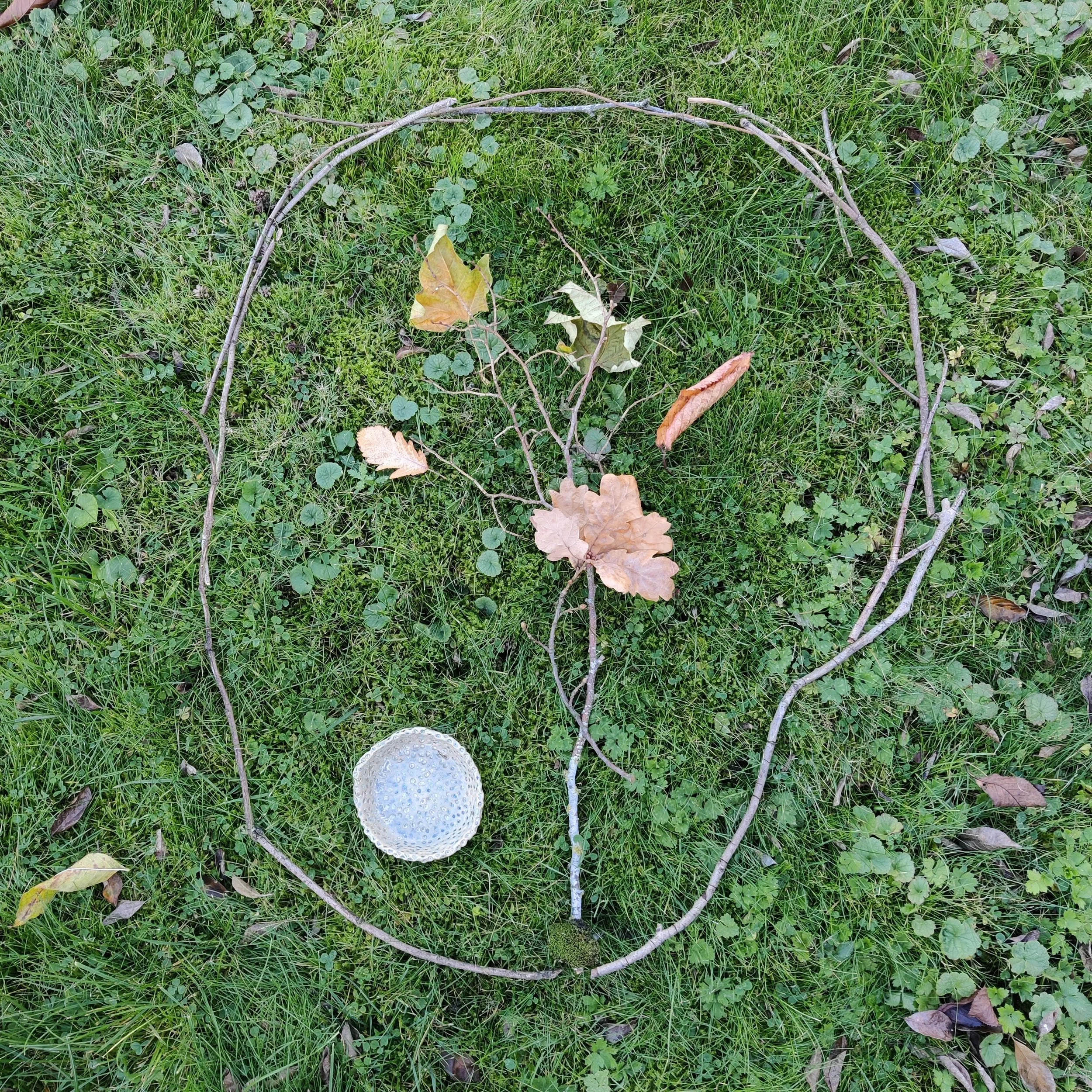Sticks, small branches, leaves and a piece of pottery arranged in a circle on grass to create a sand drawing.
