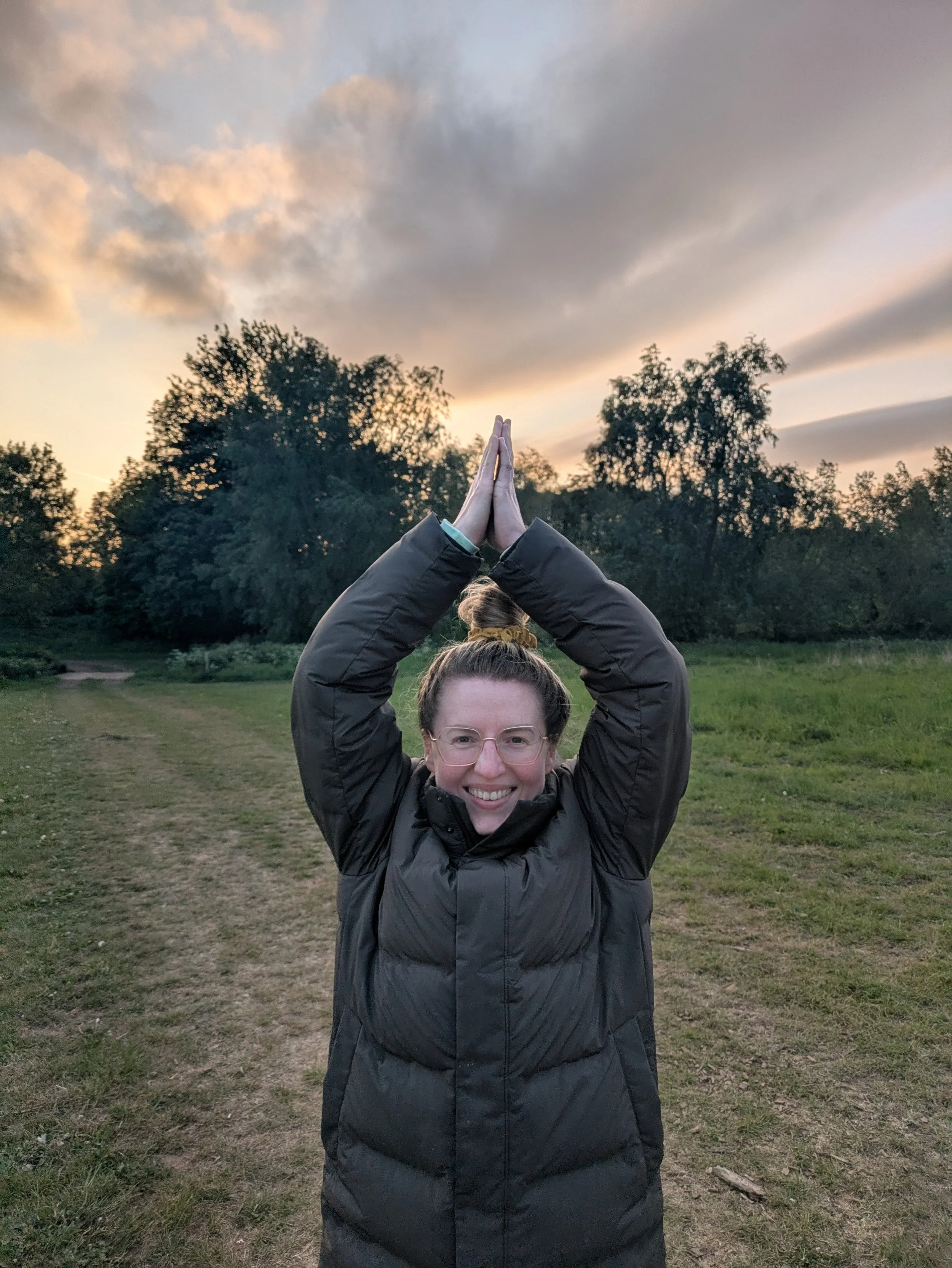 A woman wearing a green puffer coat standing outside in yoga pose with arms raised and palms of hands together above head.