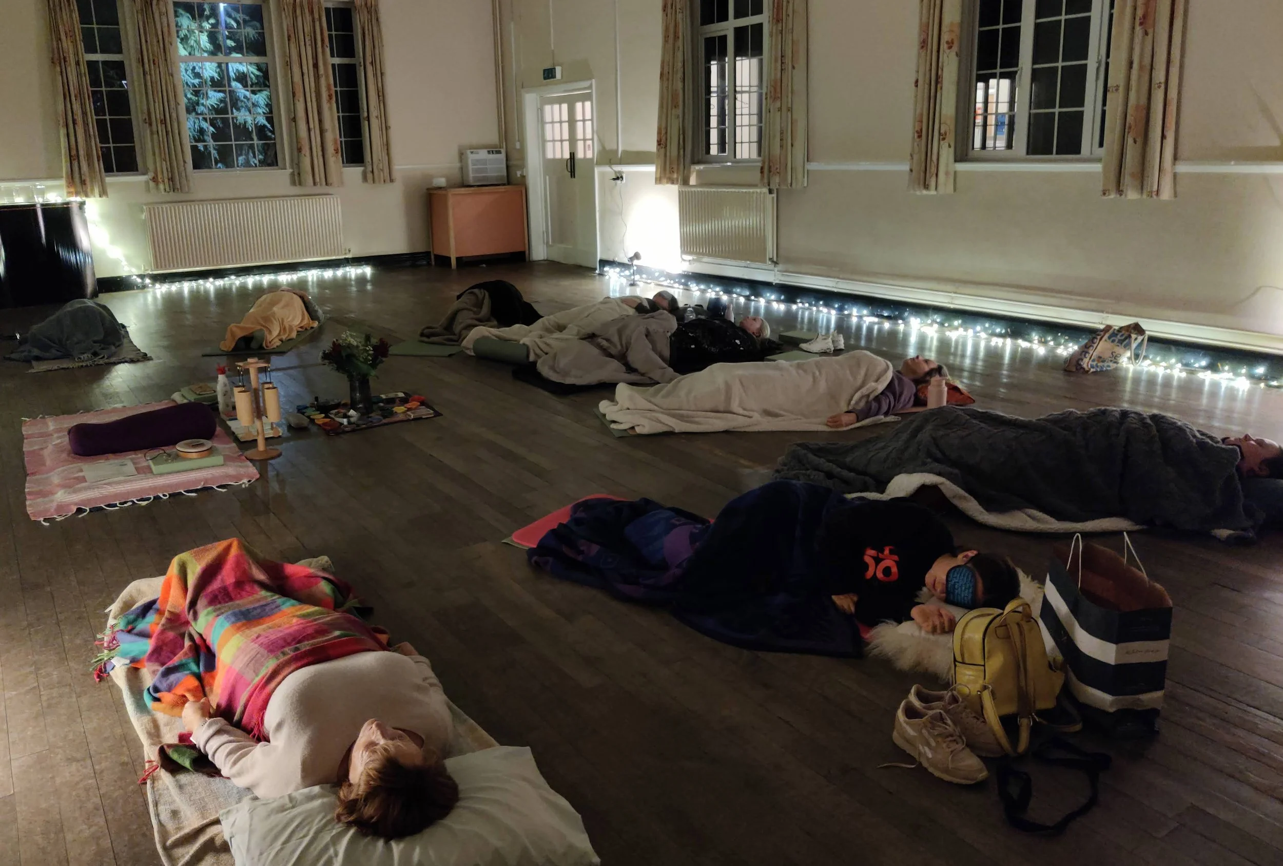 Group of people laying on mats with blankets on them in a dimly lit hall.