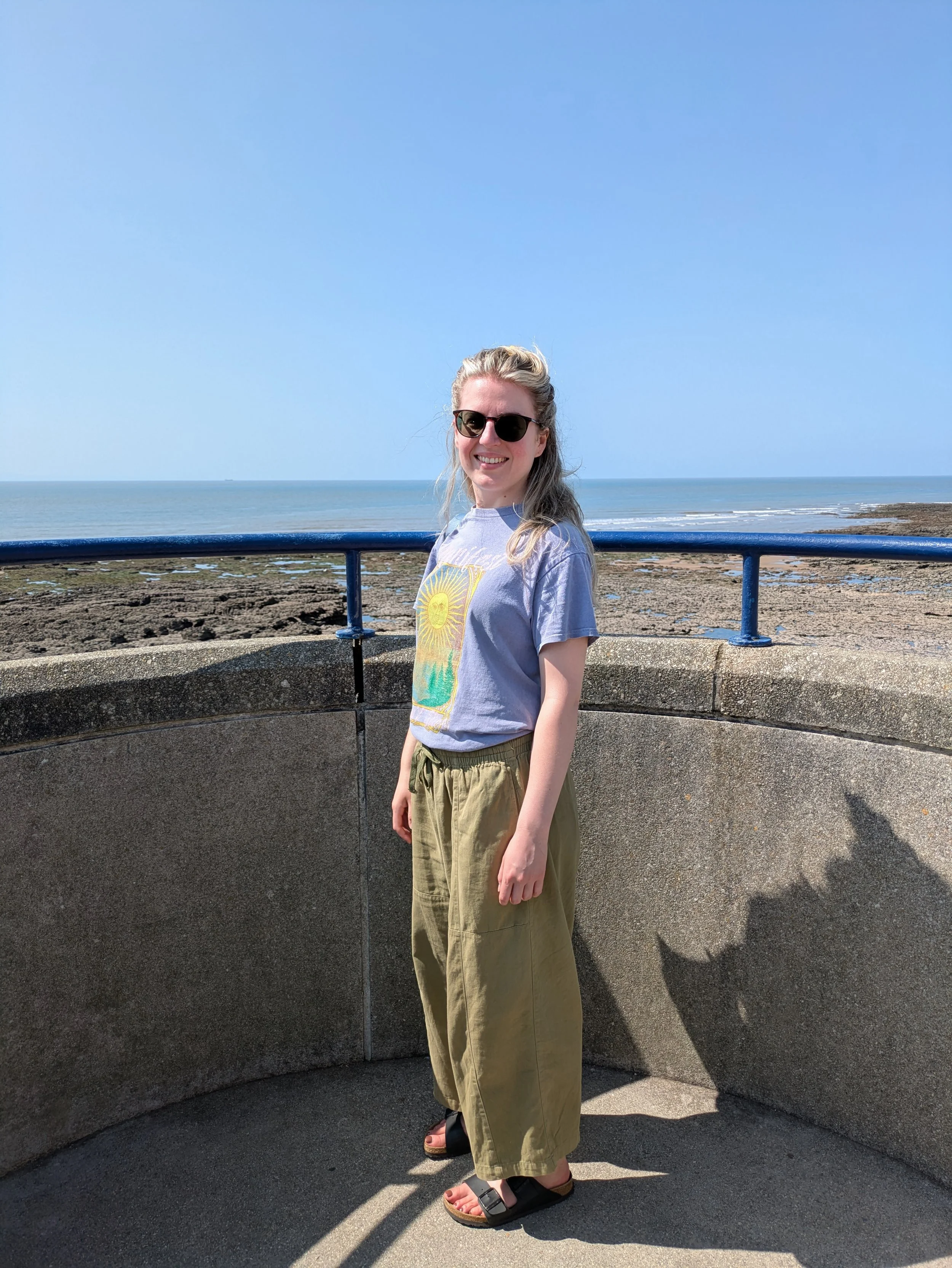 Woman standing on a concrete viewing platform in front of a sunny beach, smiling at the camera..