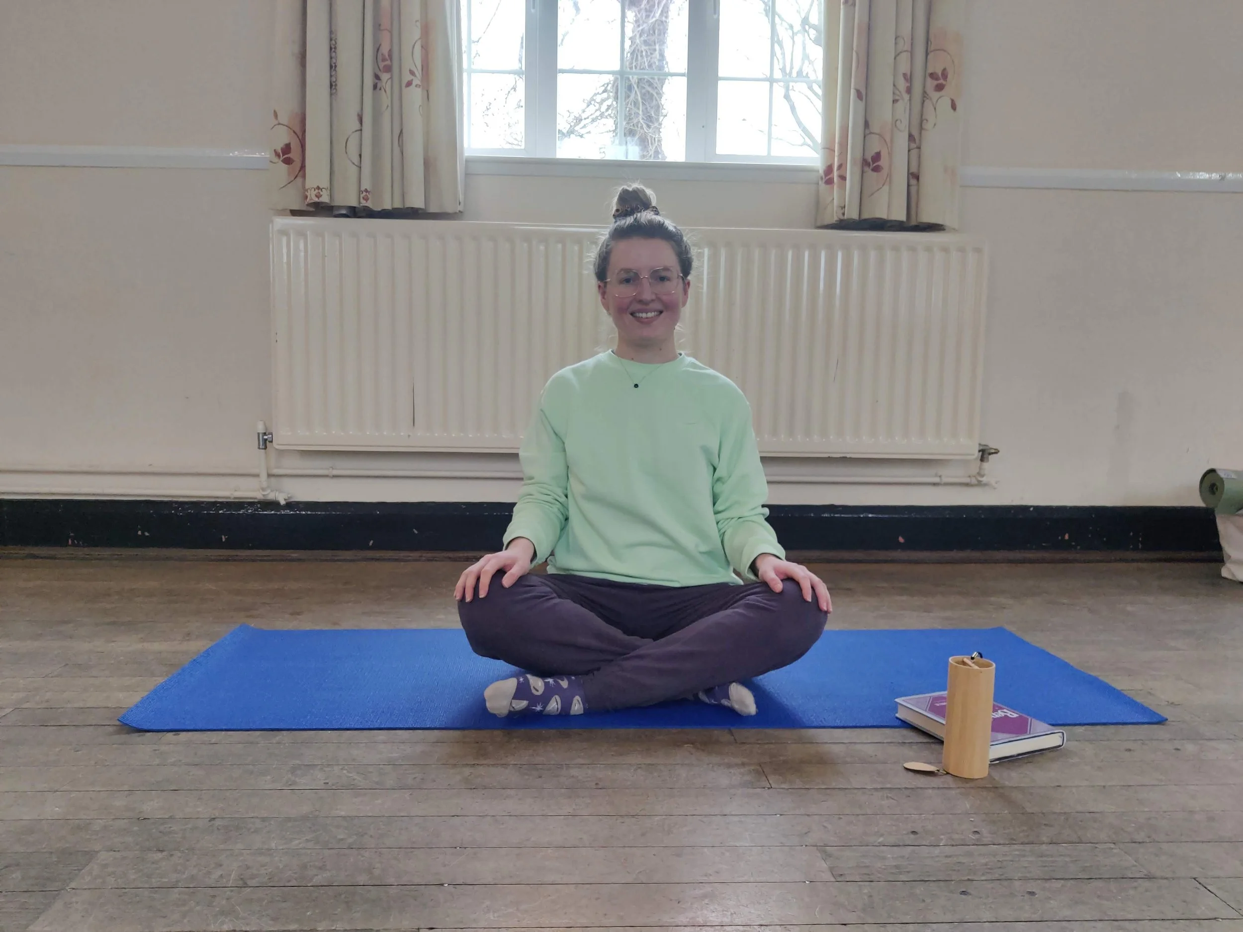 A woman sitting cross legged on a blue yoga mat gently smiling at the camera.