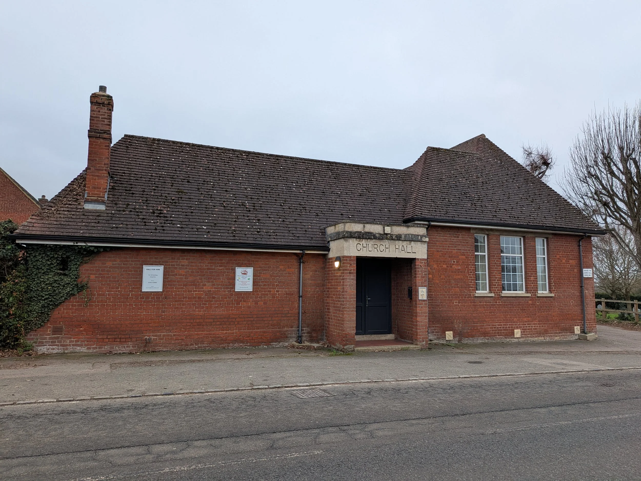 Photo of the front of All Saints Church Hall Building, which has red bricks, grey roof, a grey door in the middle and three windows on the right hand side.
