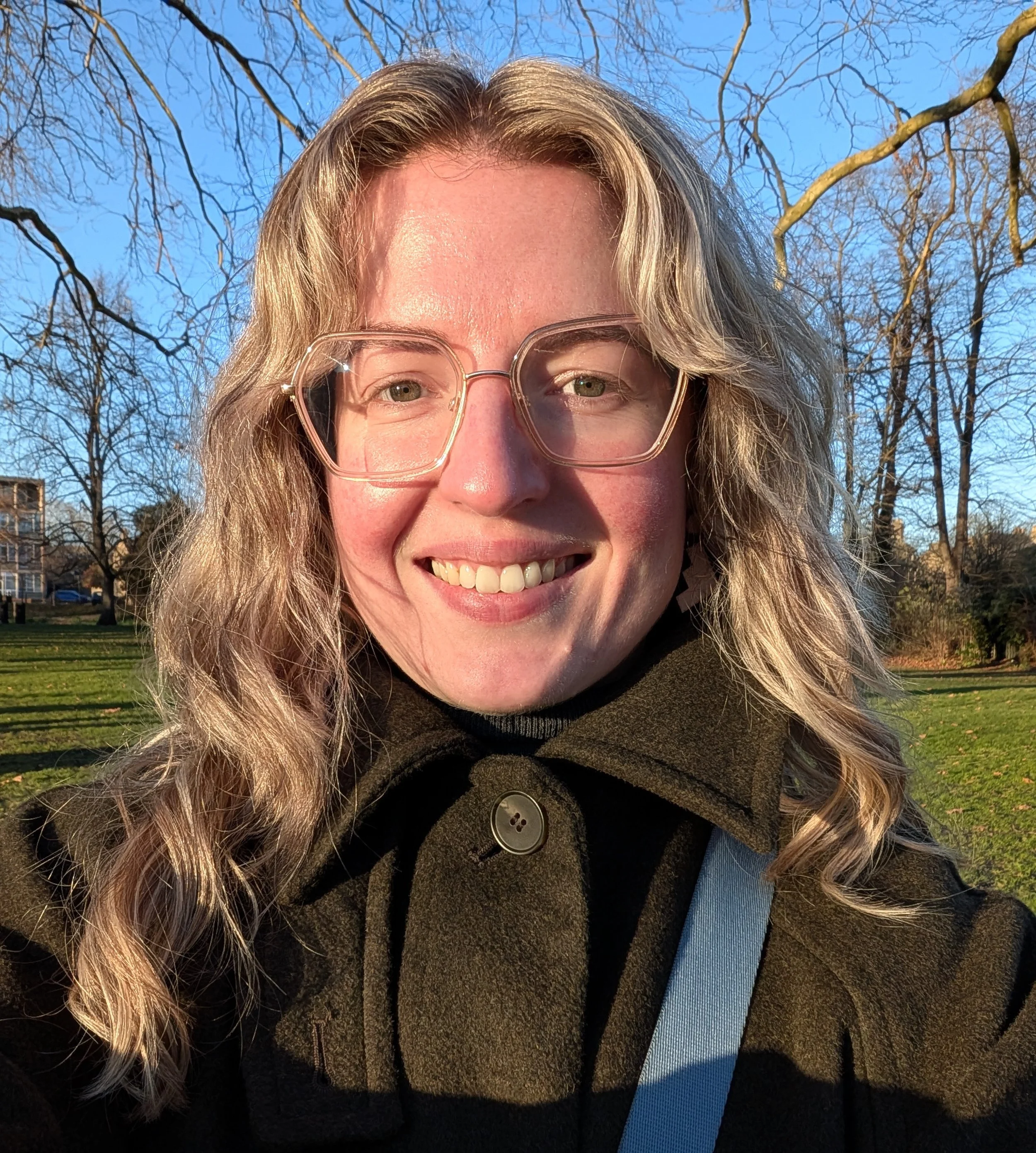Woman with blonde wavy hair and glasses in a green coat with blue sky, tree branches and grass behind her, gently smiling at the camera.