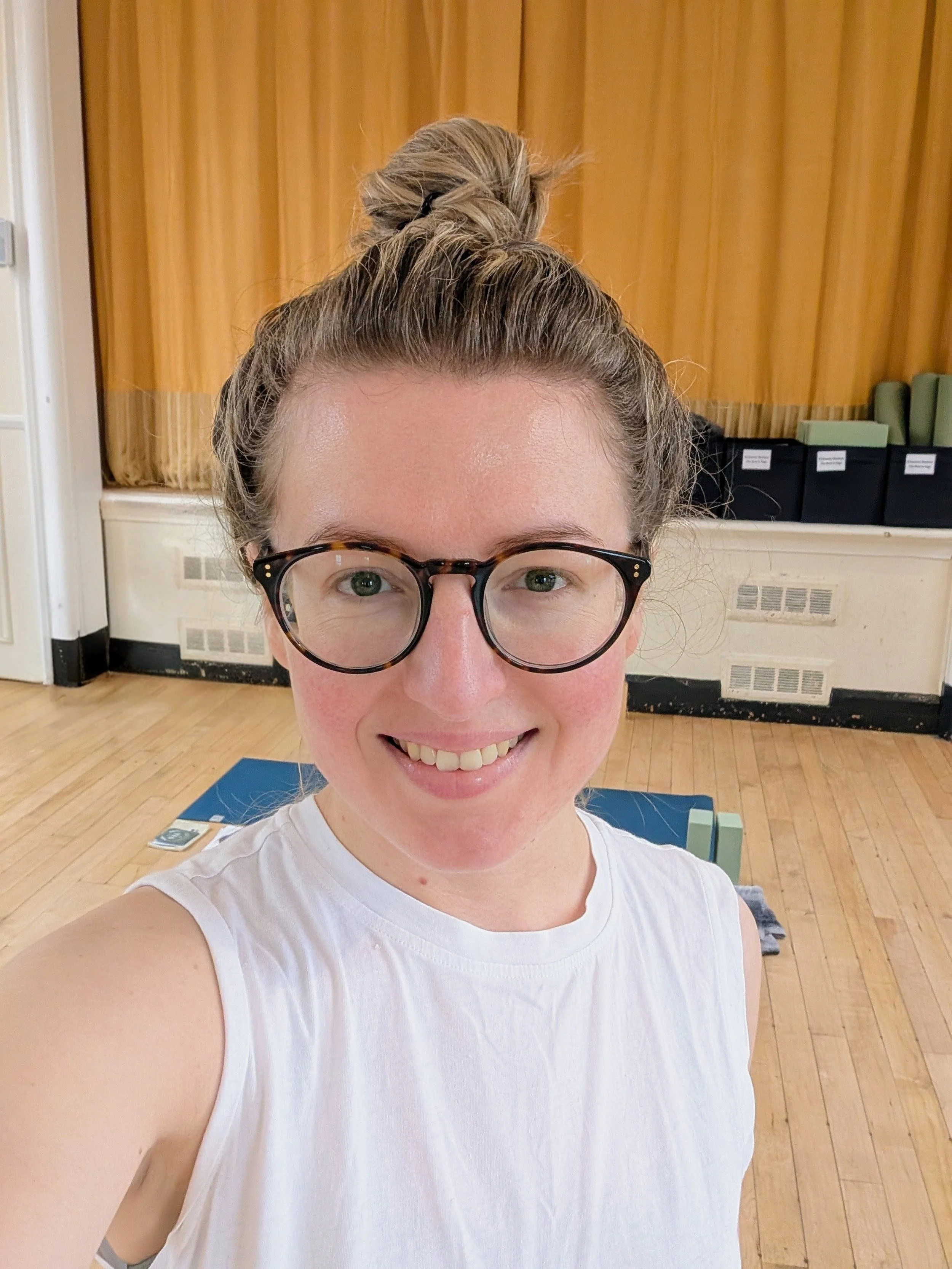 A woman with glasses and hair in a bun smiling at the camero standing in front of a yoga mat ready for a yoga class in Clifton, Bedfordshire.