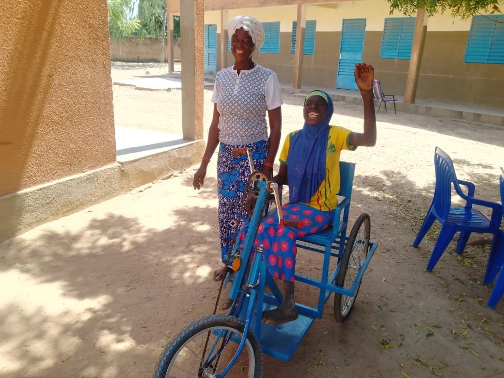 Zwei lachende Frauen, eine davon sitzend im Rollstuhl, die andere steht neben ihr, im Schatten. Sie tragen bunte Kleidung und scheinen glücklich zu sein, im Hintergrund eine Schulhof-Szene.