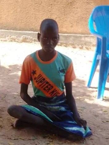 Junge sitzt im Sand, trägt bunte T-Shirt, im Hintergrund blaue Stühle.