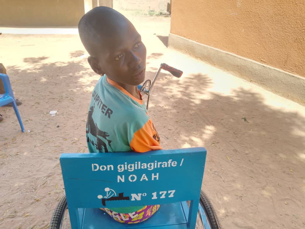 Ein Junge im Rollstuhl sitzt auf einem blauen Stuhl mit einem Schild, auf dem steht: 'Don gigilagira / NOAH, N° 177'.