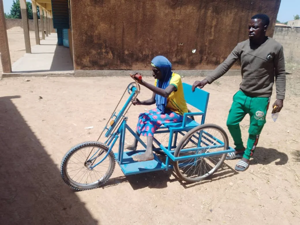 Eine Frau sitzt auf einem blauen Rollstuhl, der wie ein Fahrrad aussieht, während ein Mann sie schiebt.