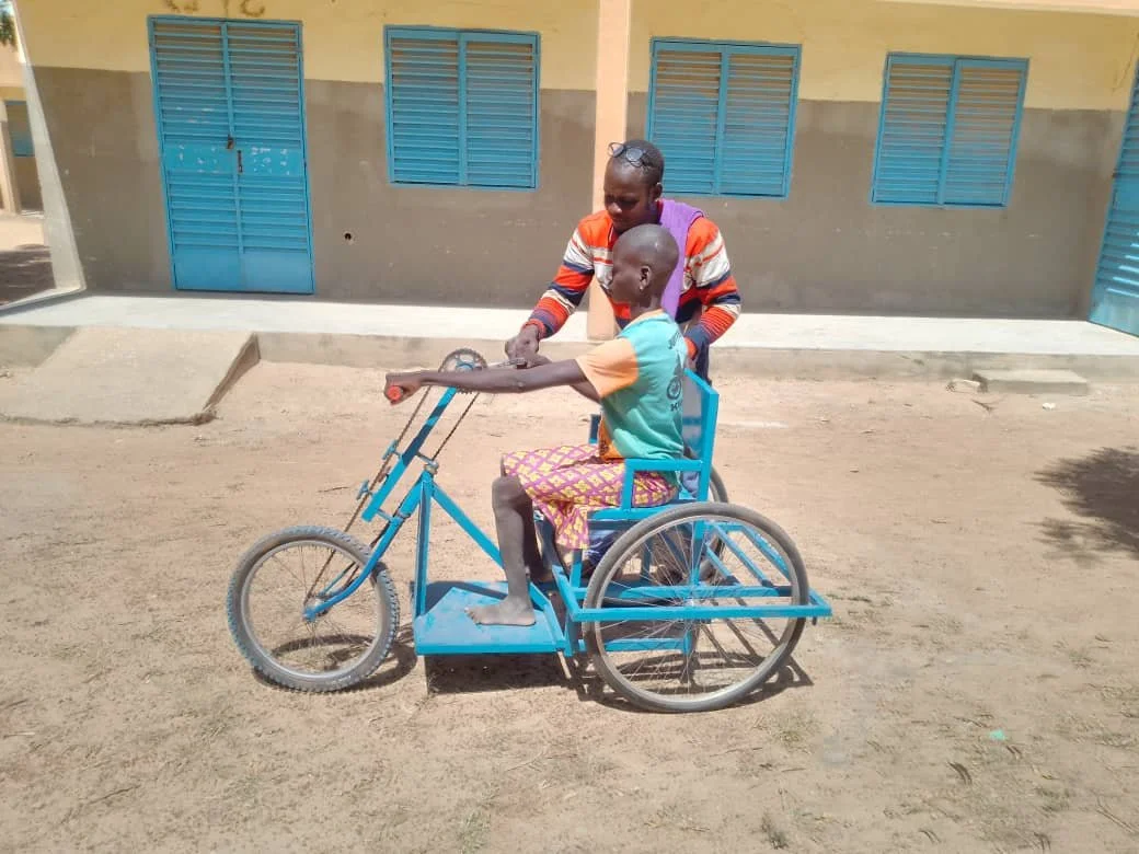 Ein Junge sitzt in einem schwarzen Rollstuhl mit blauer Metallrahmen, während eine andere Person, vermutlich ein Erwachsener, ihm beim Fahren des Rollstuhls hilft. Der Hintergrund zeigt ein Gebäude mit blauen Fensterläden und einer grauen unteren Wan