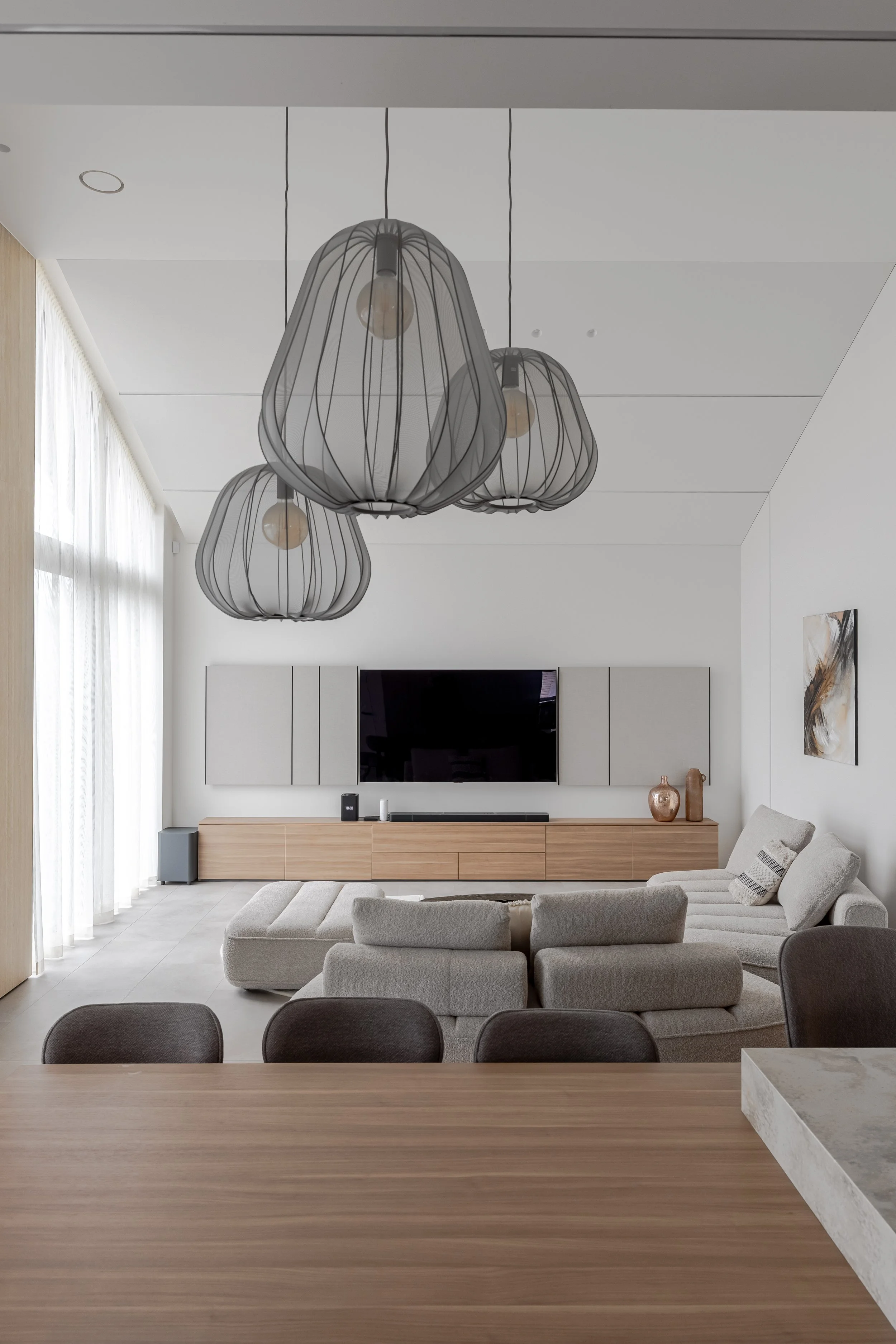 Modern living room with a beige sectional sofa, wall-mounted TV, wooden cabinet, abstract artwork, pendant lights, and sheer curtains.