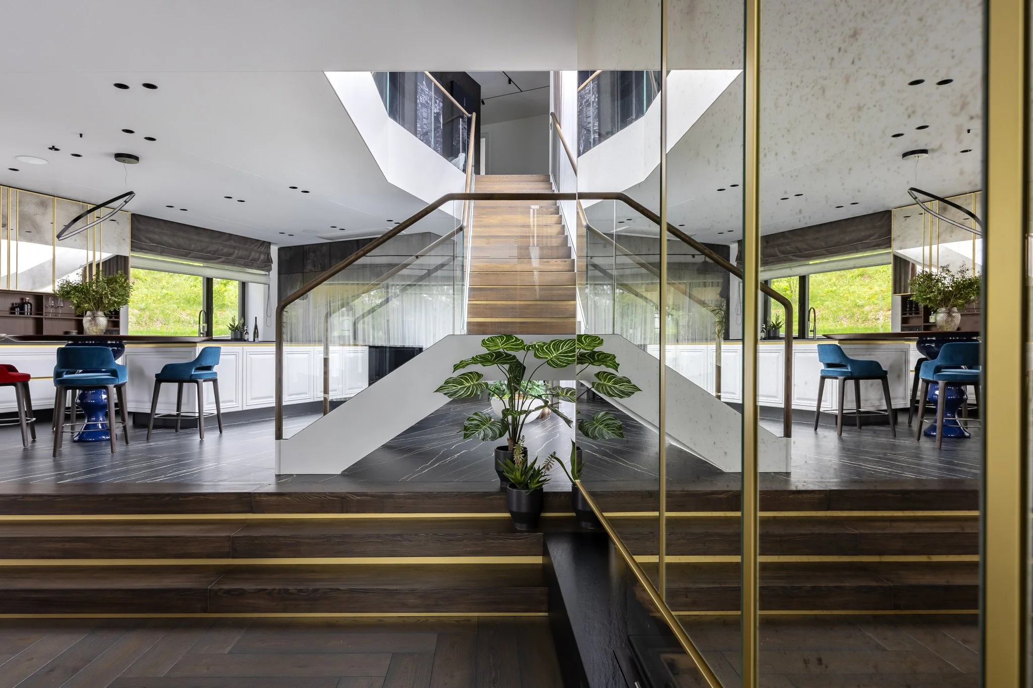 Modern interior with staircase, glass railings, blue chairs, large windows, green plants, and wooden accents.