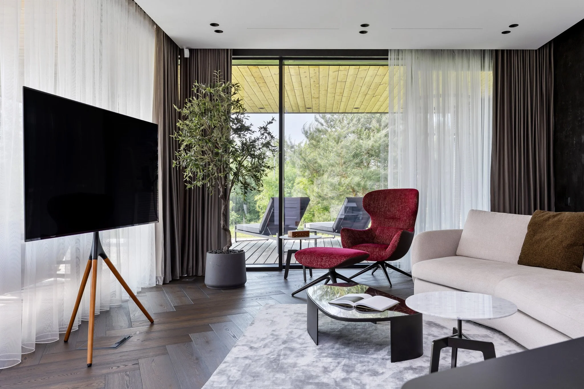Modern living room with a large flat-screen TV on a wooden stand, a beige sofa, a red armchair with an ottoman, a coffee table with an open book, a potted indoor tree, and sliding glass doors leading to a balcony with outdoor chairs and greenery outside.