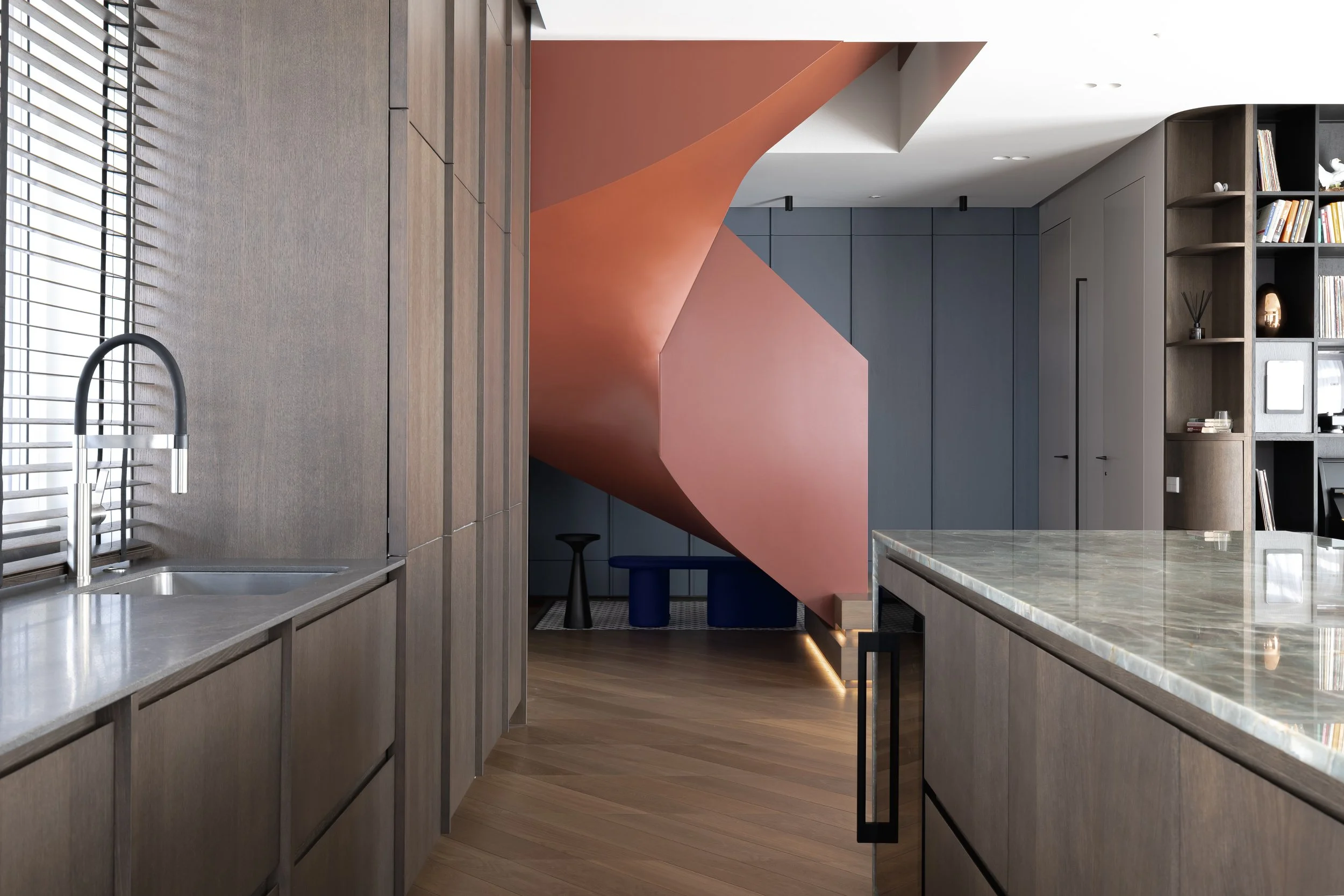Modern kitchen with wooden cabinets, marble countertop, window with blinds, gray walls, and a pink spiral staircase in the background.