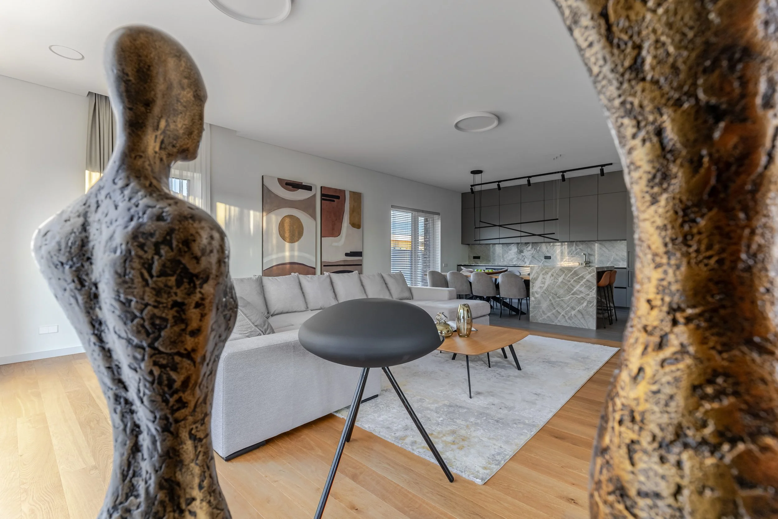 Modern living room with a white sectional sofa, abstract artwork on the wall, and a kitchen with gray cabinets and a marble island in the background. The photo is taken through a frame of two textured, brown sculptures.