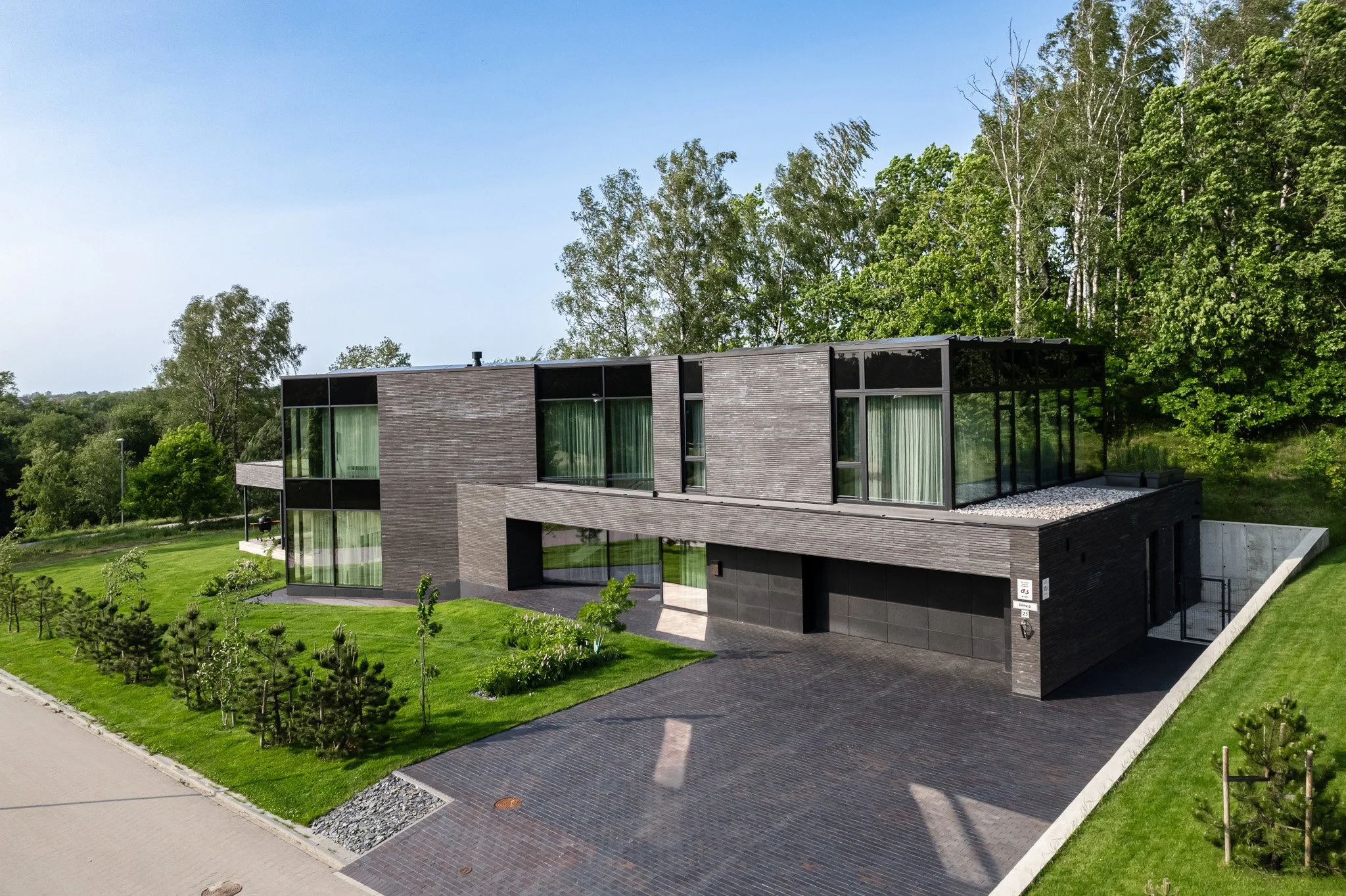 Modern, two-story house with large glass windows, dark brick exterior, surrounded by green lawn and trees, situated on a paved driveway.