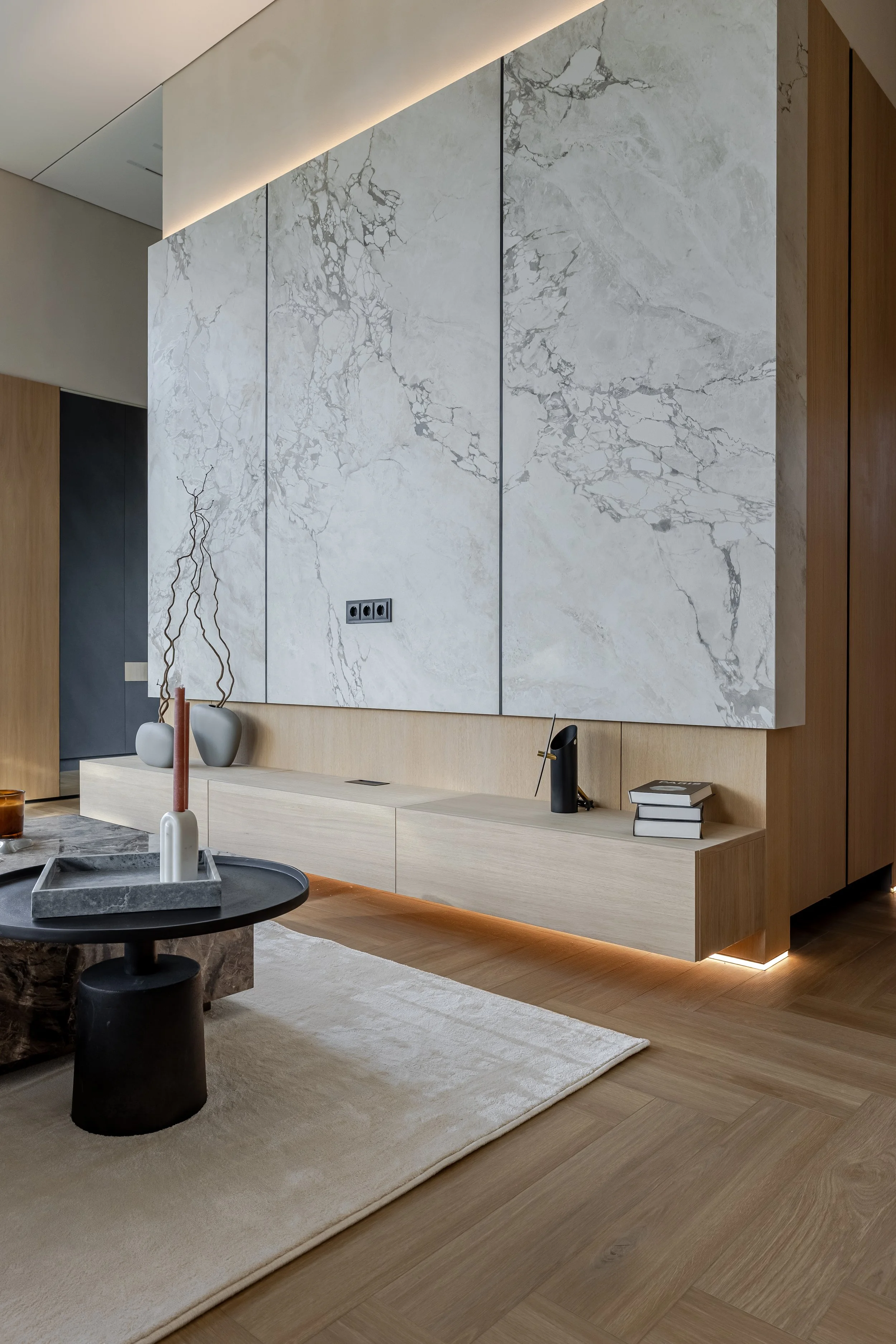 Modern living room corner featuring a large white marble wall with gray veining, a low wooden sideboard with minimal decor including vases and books, a circular coffee table with a tray and candles, and a beige rug on wooden flooring.