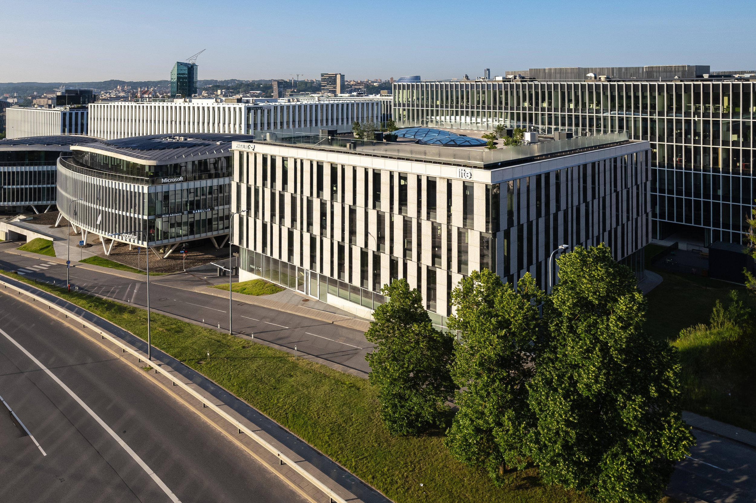 Modern office buildings with glass facades, trees along the sidewalk, and a multi-lane road in the foreground.
