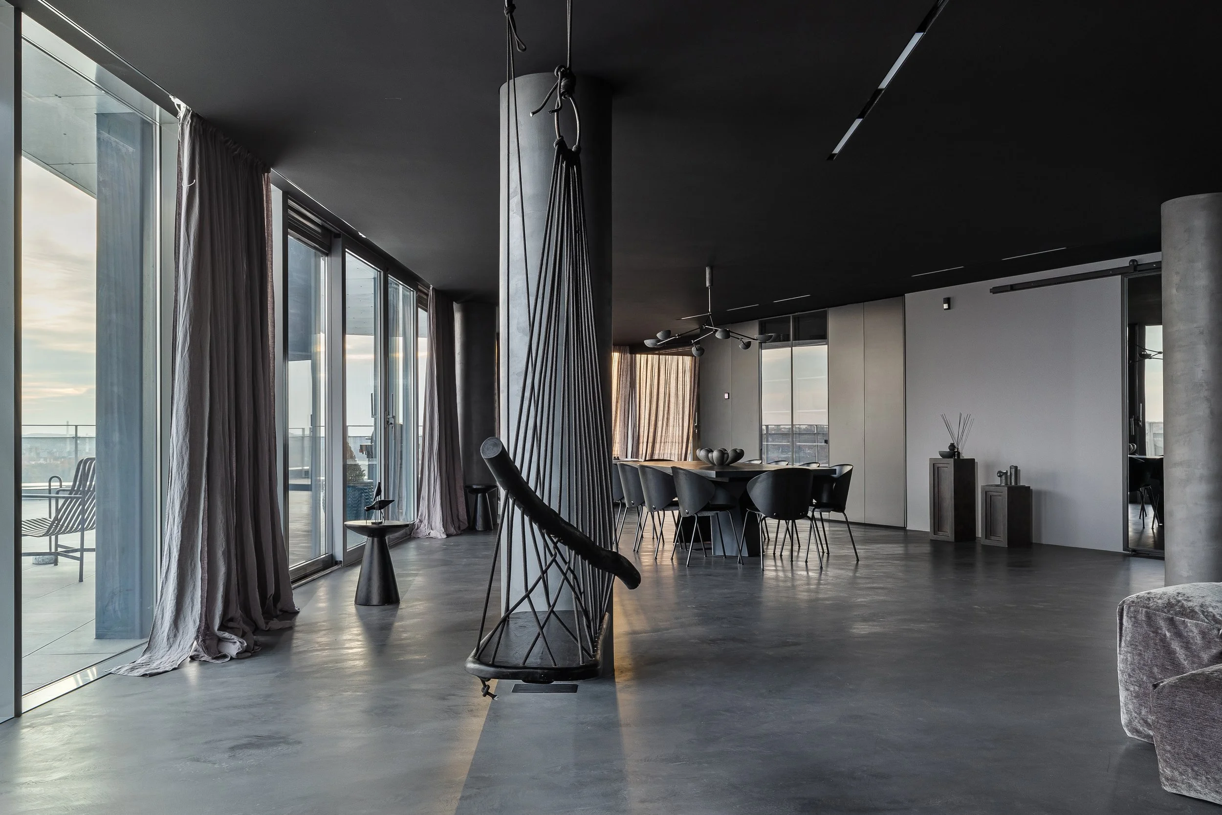 Modern living room with large windows, a dining table with black chairs, a black swing chair, and minimalistic decor.