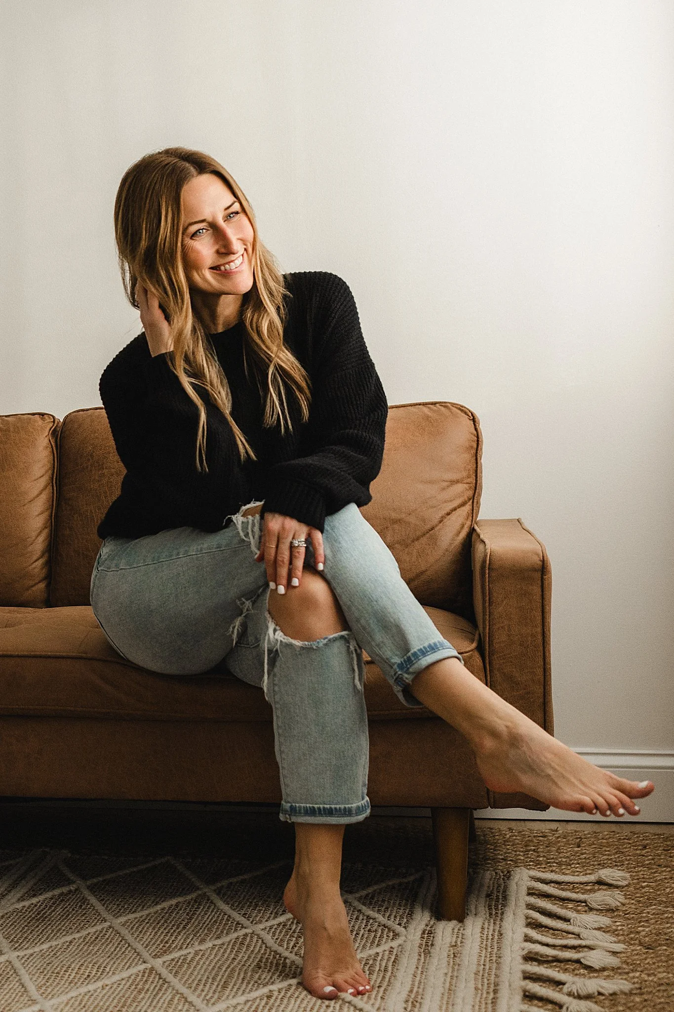 A woman named Marie Lopez, author of The Summer We Kissed at Blackbird, with long wavy hair smiling, sitting on a brown couch, wearing a black sweater and ripped jeans, with one leg crossed over the other, in a room with a plain wall.