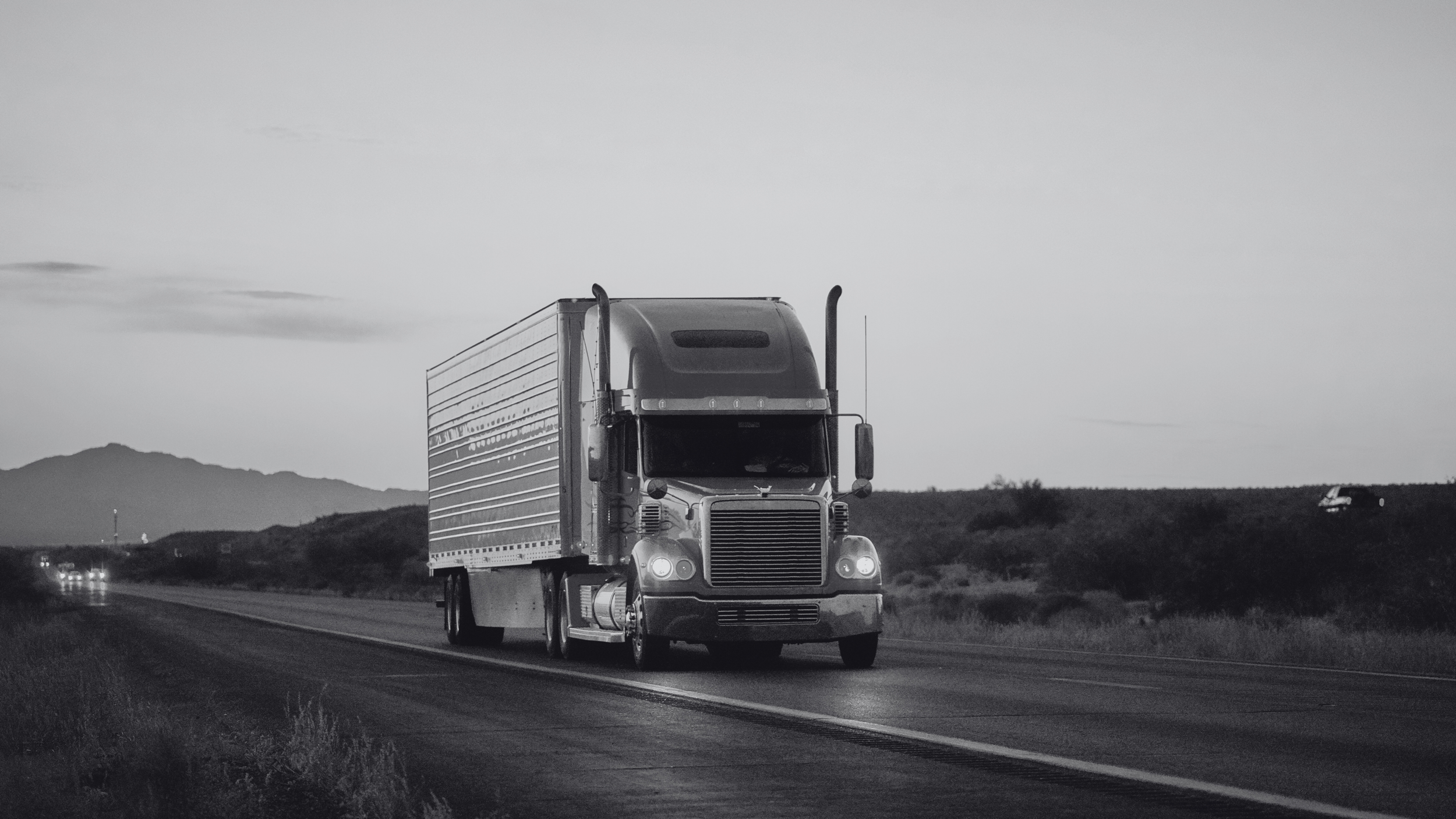 A semi-truck driving on an open road with mountains and a cloudy sky in the background, moving music from Scandinavia to cinematic advising companies around the world.