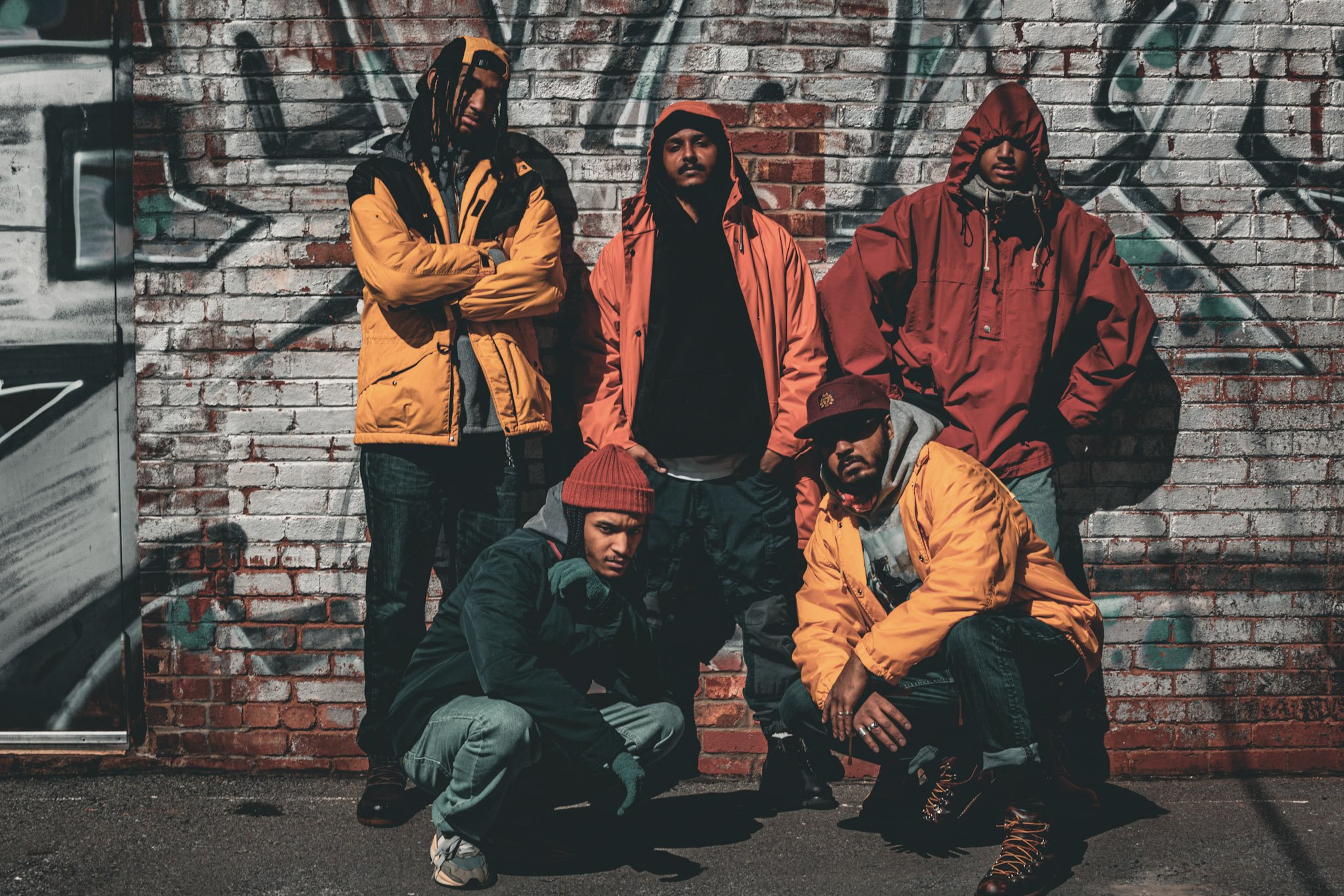 Clan Supreme members photoshoot against a graffiti-covered brick wall, with some standing and others crouching, dressed in colorful streetwear jackets and casual clothing.