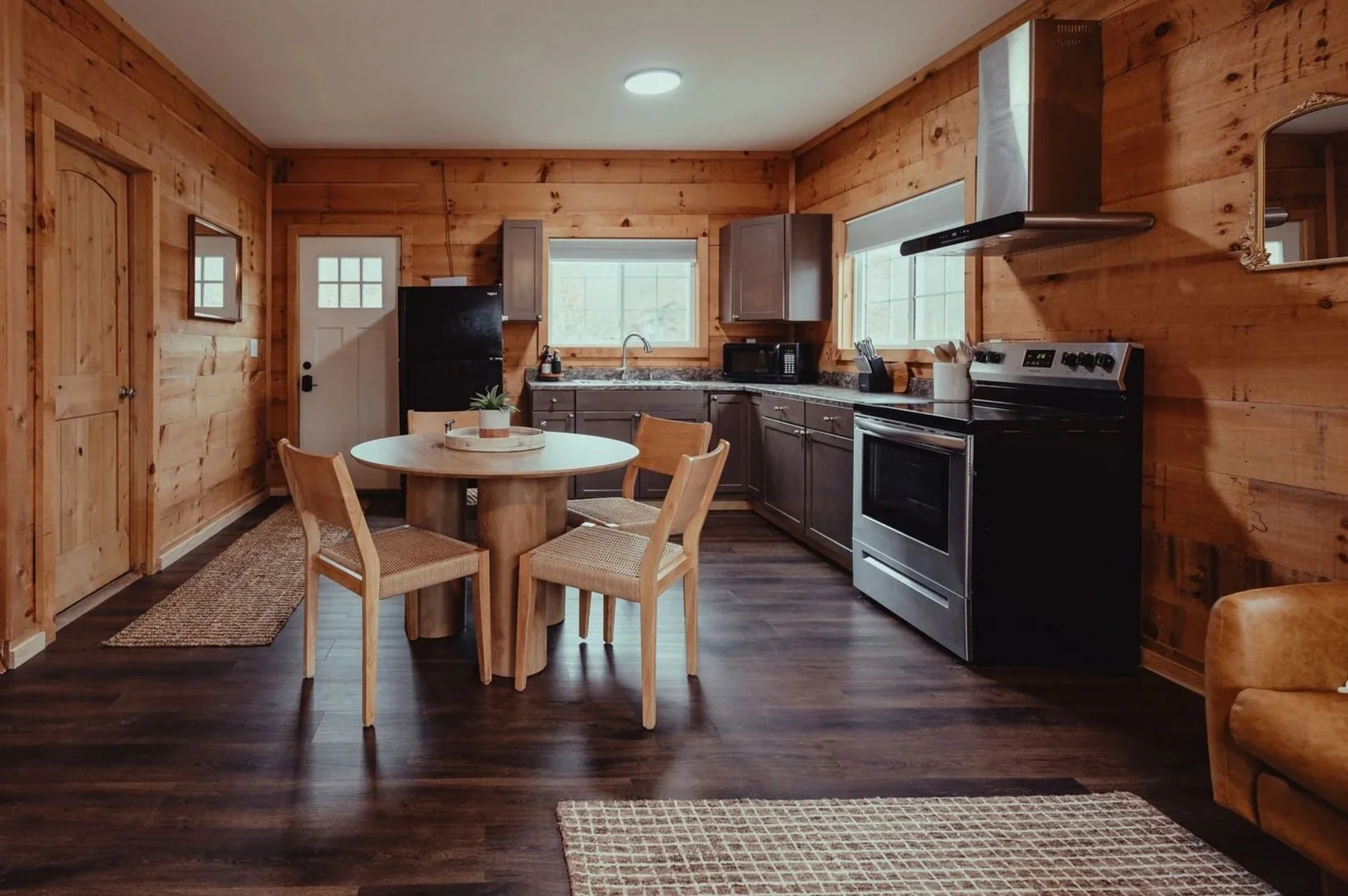 Interior of a rustic wooden kitchen with a round wooden dining table, four chairs, dark gray cabinets, a black refrigerator, stainless steel oven, microwave, wooden walls, and windows letting in natural light.