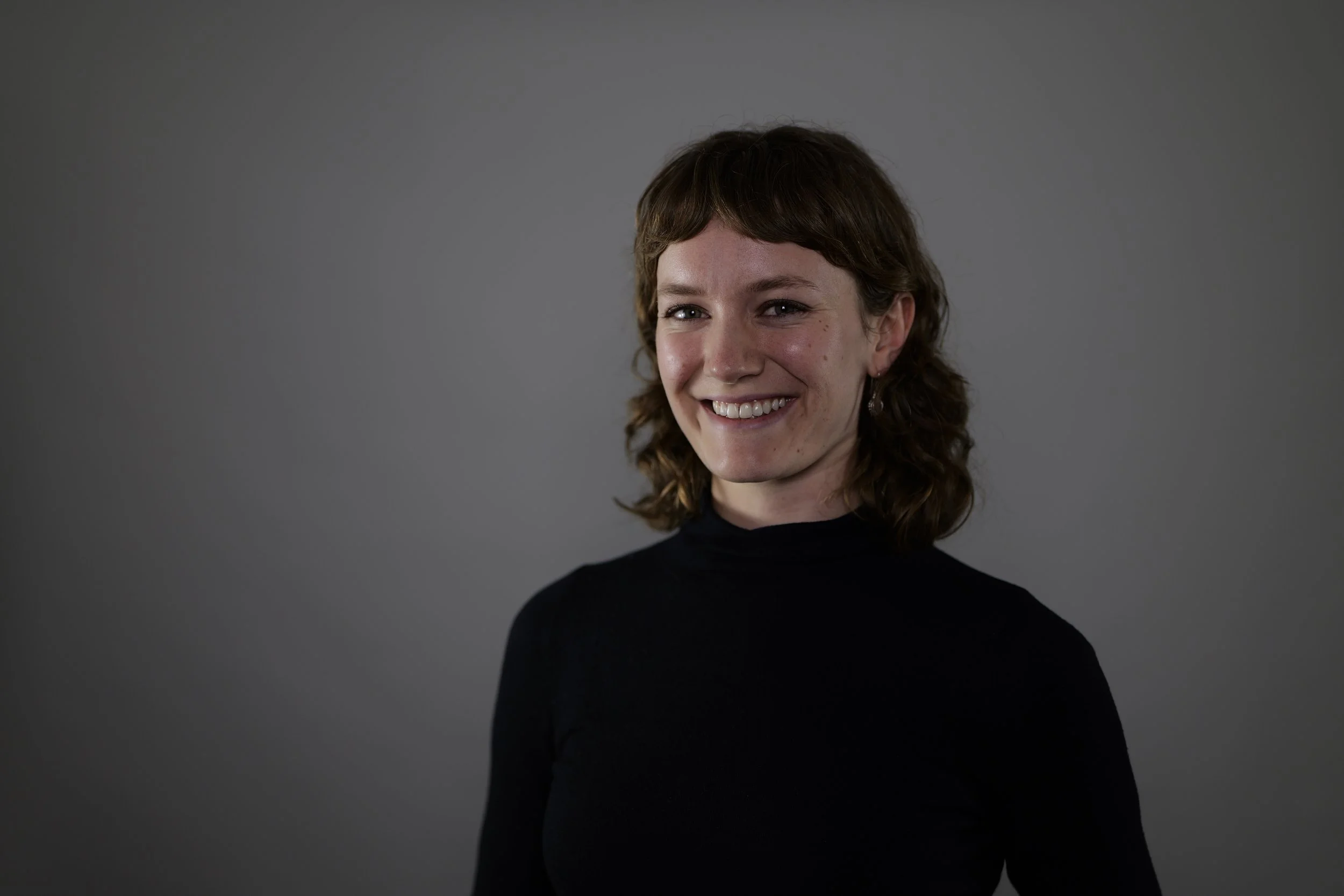 A woman with shoulder-length brown hair, smiling, wearing a black turtleneck, standing against a plain gray background.