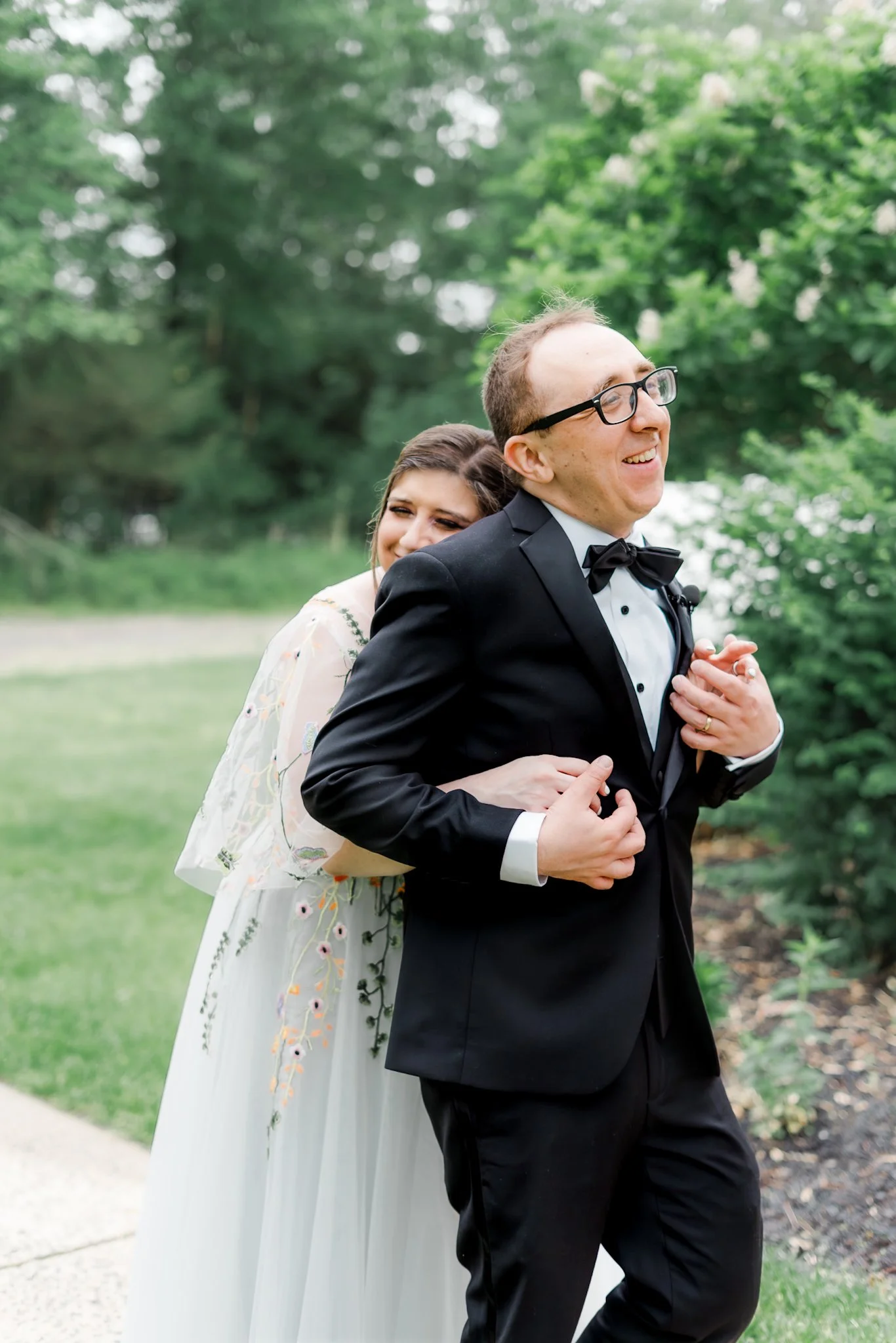 A couple dressed in formal wedding attire, smiling and embracing outdoors with green trees in the background.