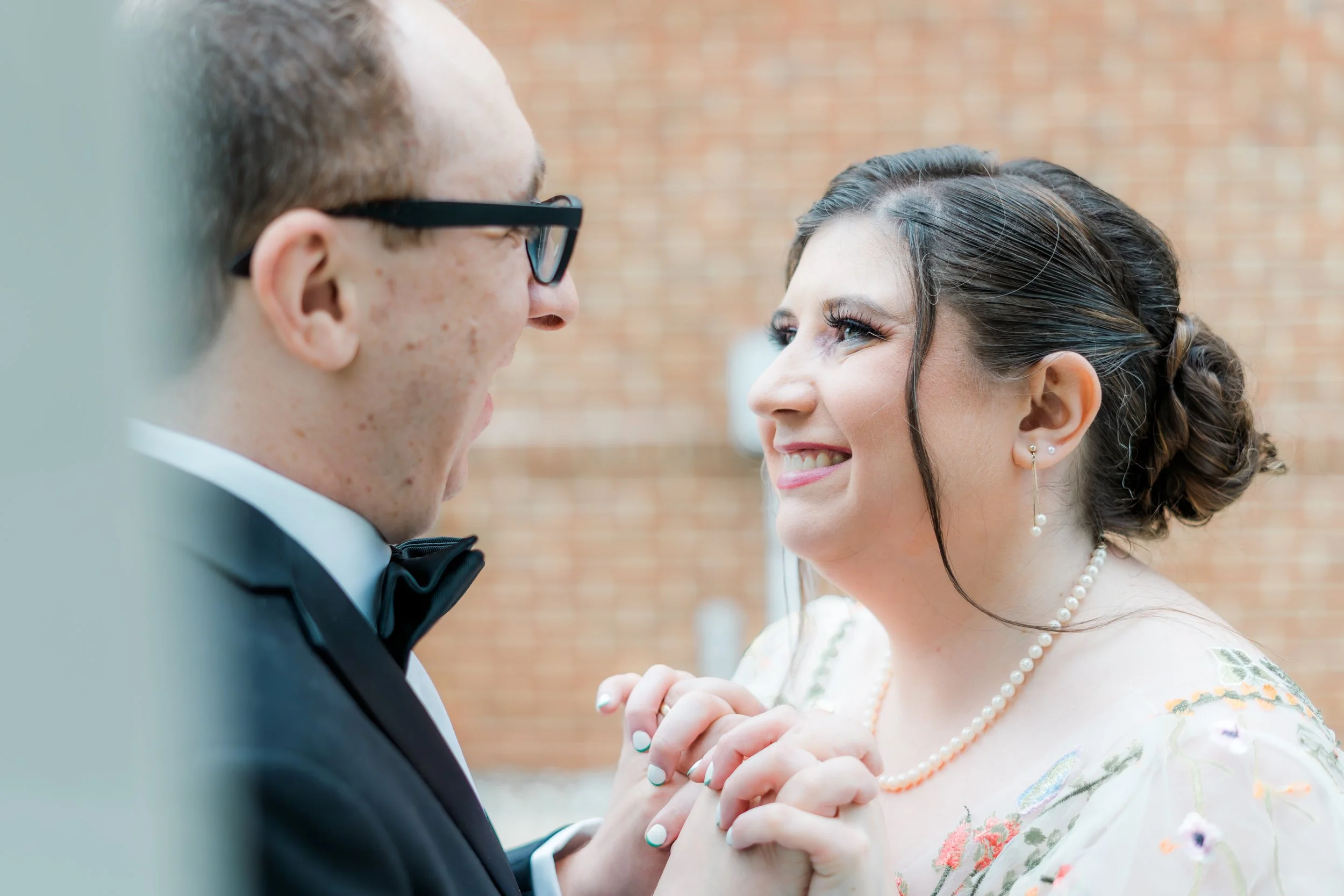A bride and groom holding hands and smiling at each other, dressed in wedding attire, with a brick wall in the background.