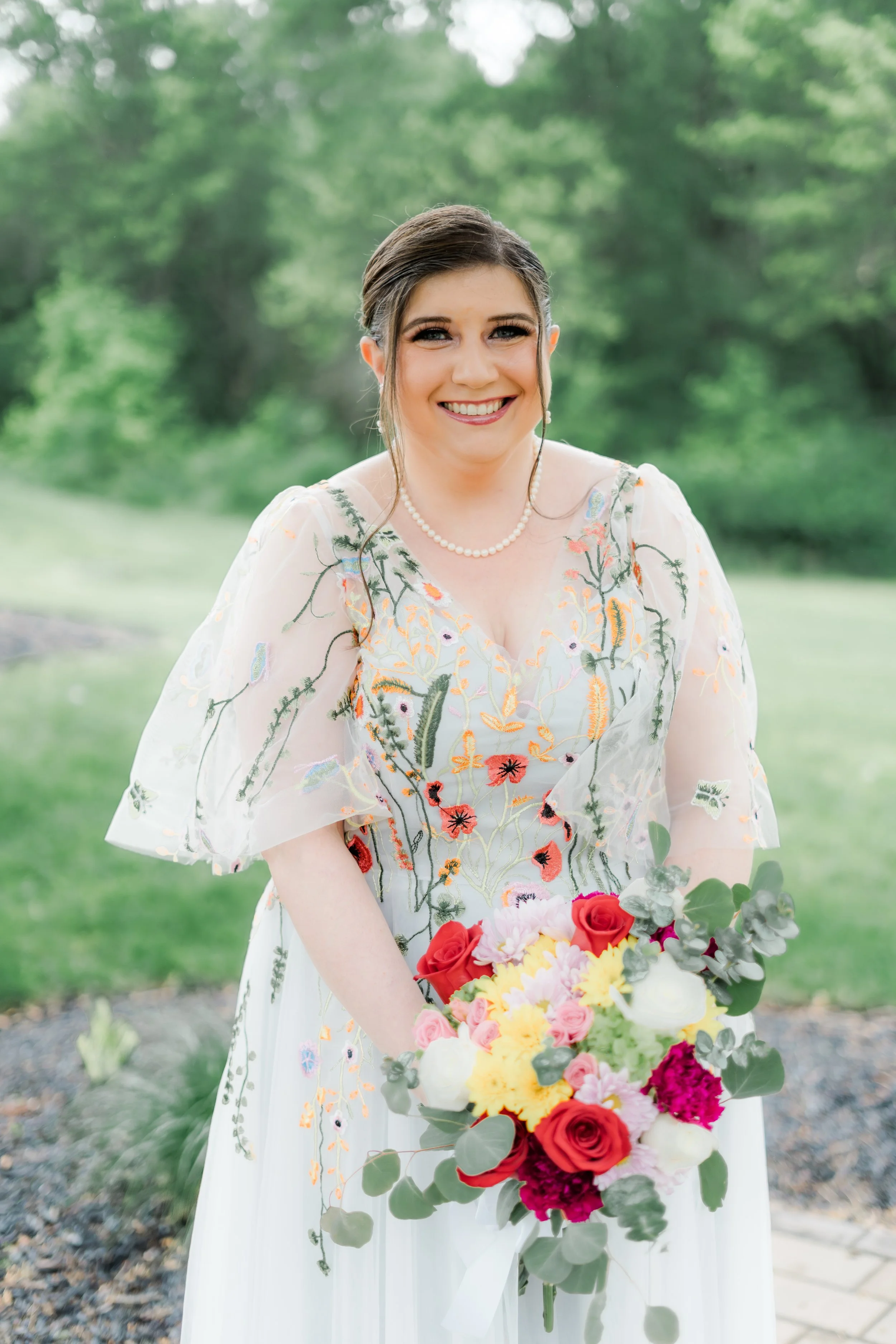 A smiling woman in a white dress with floral embroidery holds a bouquet of colorful flowers outdoors with blurred green trees in the background.