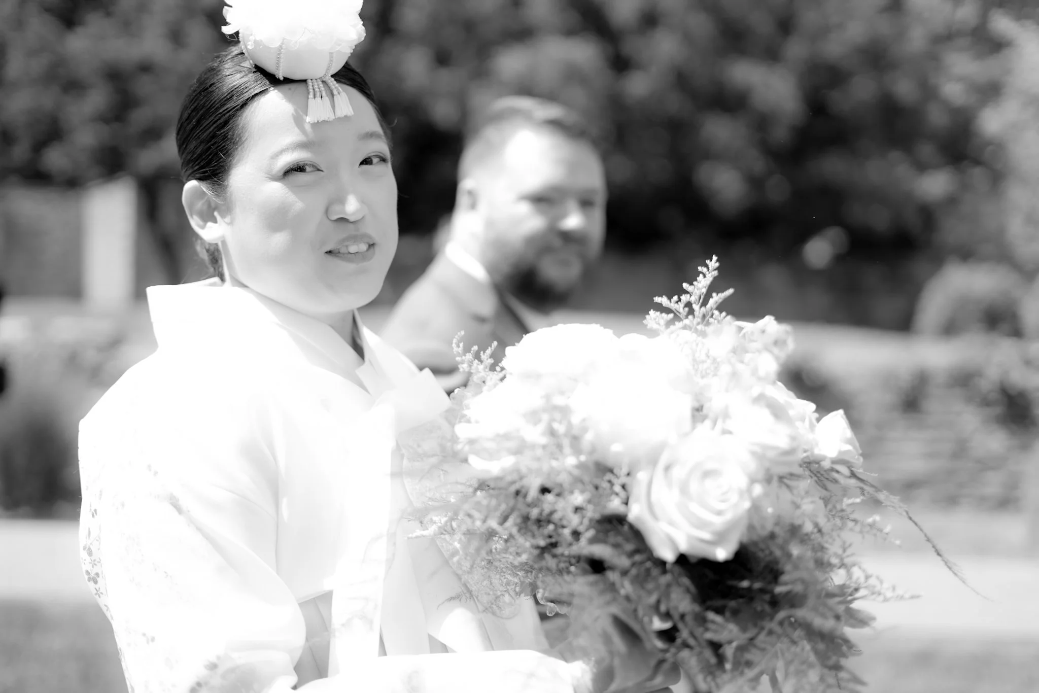 Black and white photo of a woman in traditional Japanese attire holding a bouquet of flowers, with a man smiling in the background. The woman is wearing a headpiece and has a gentle expression.