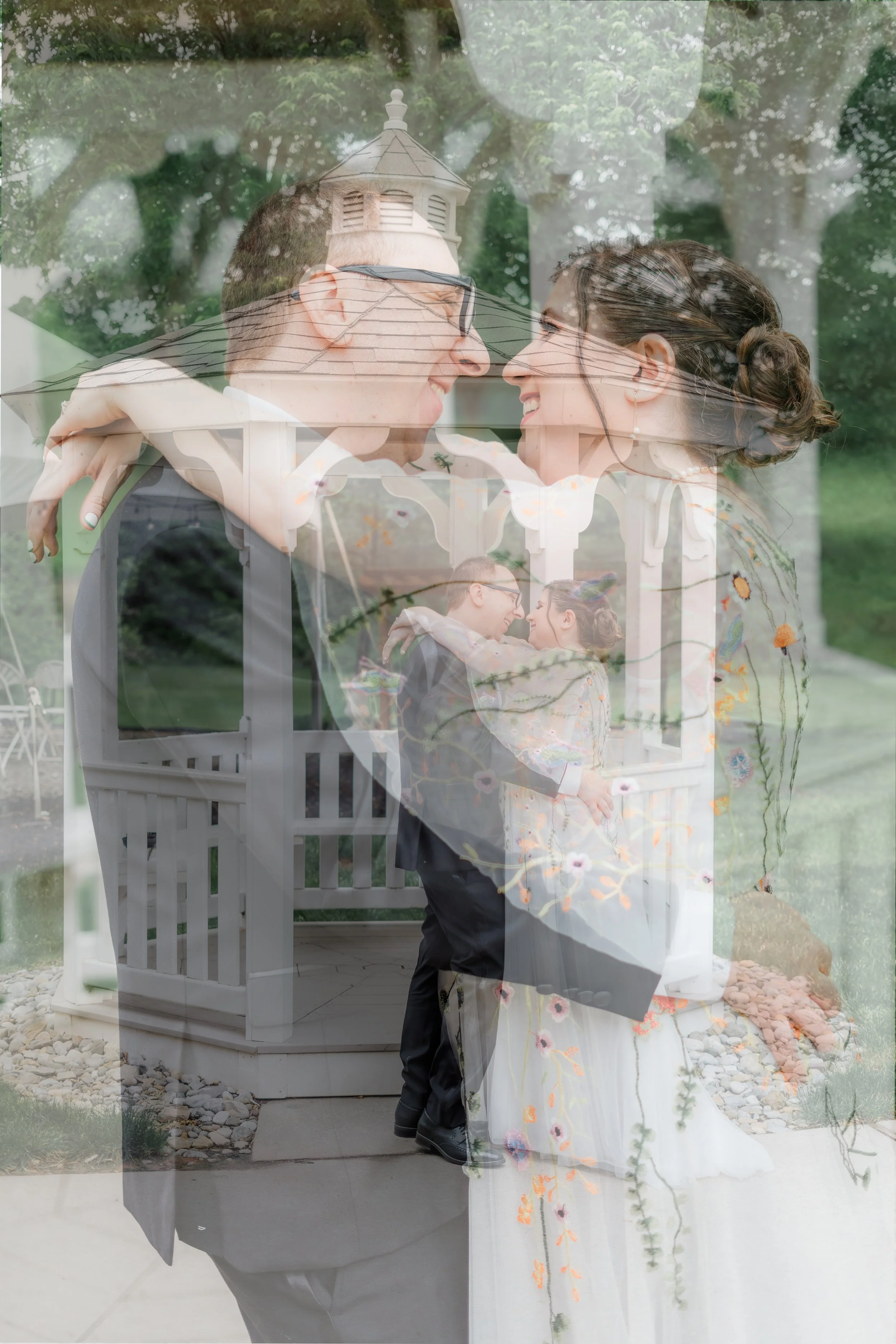A couple is embracing and smiling at each other in front of a gazebo, with the woman's wedding dress and the man's suit visible. The photo is taken through a window, creating a layered reflection of trees and sky.