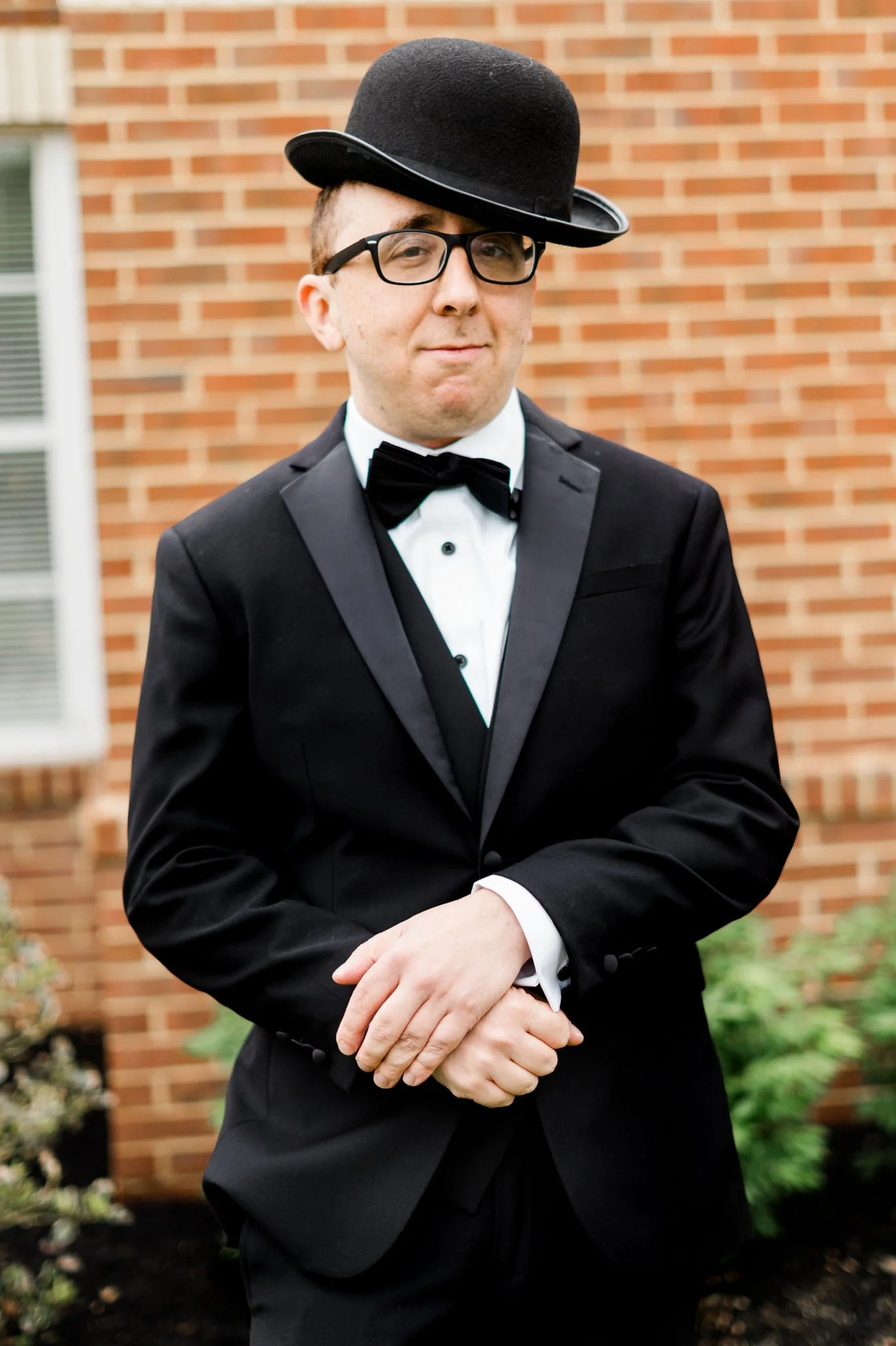 A man dressed in a tuxedo with a bow tie and glasses, wearing a bowler hat tilted sideways, standing outdoors in front of a brick building.